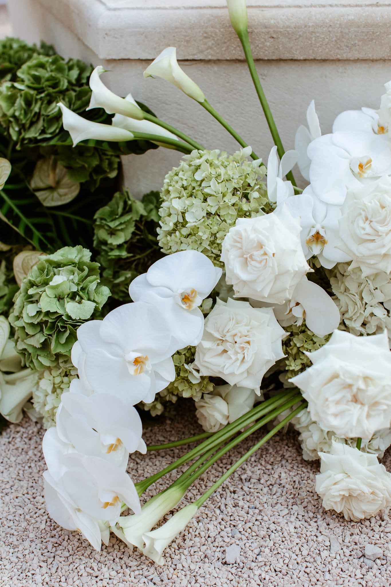 Close-up of white orchids, ivory garden roses, calla lilies, and sage-green hydrangeas on pale gravel