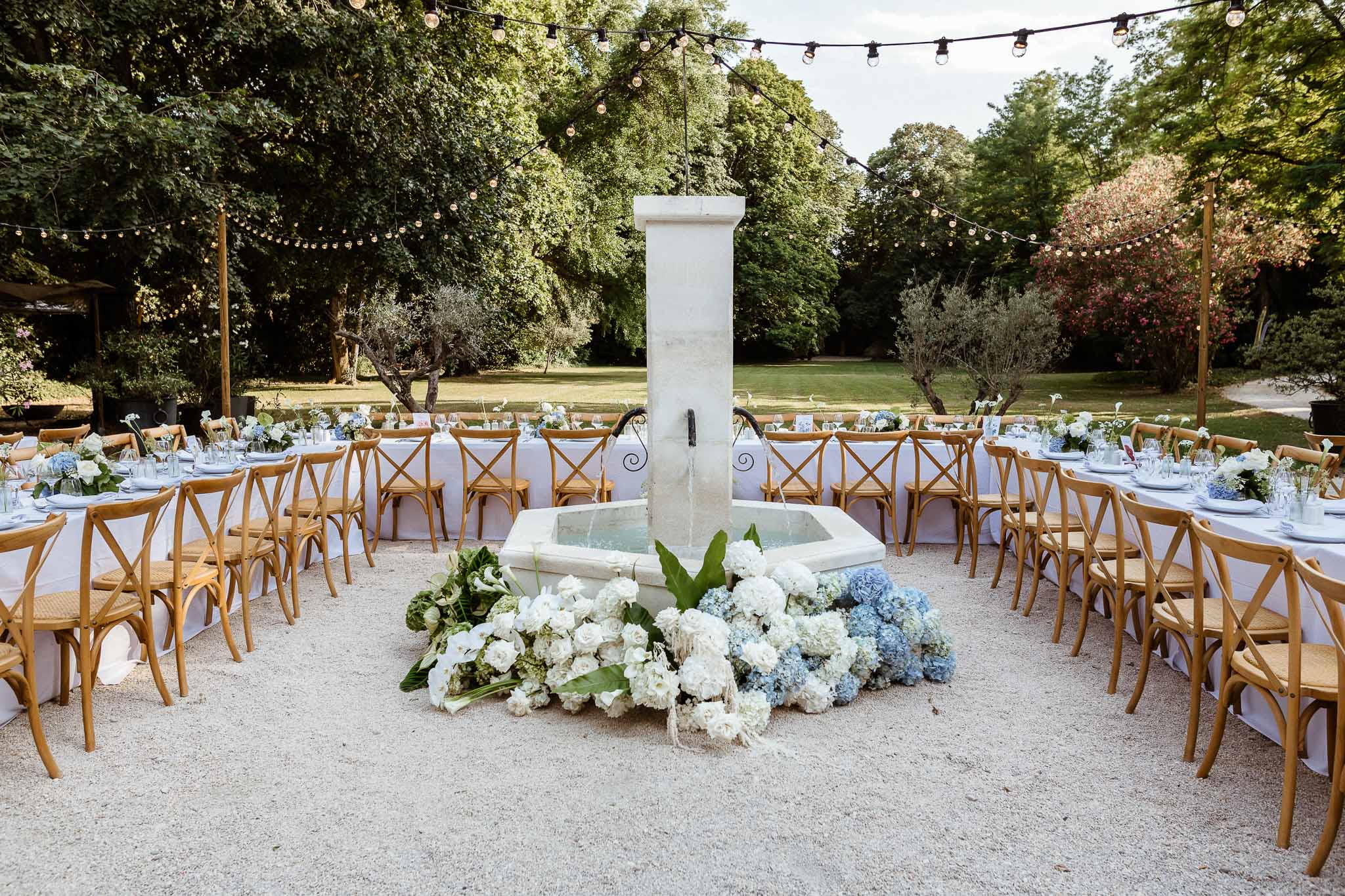 Outdoor reception U-shaped tables around stone fountain with white and blue hydrangeas and Edison bulb string lights