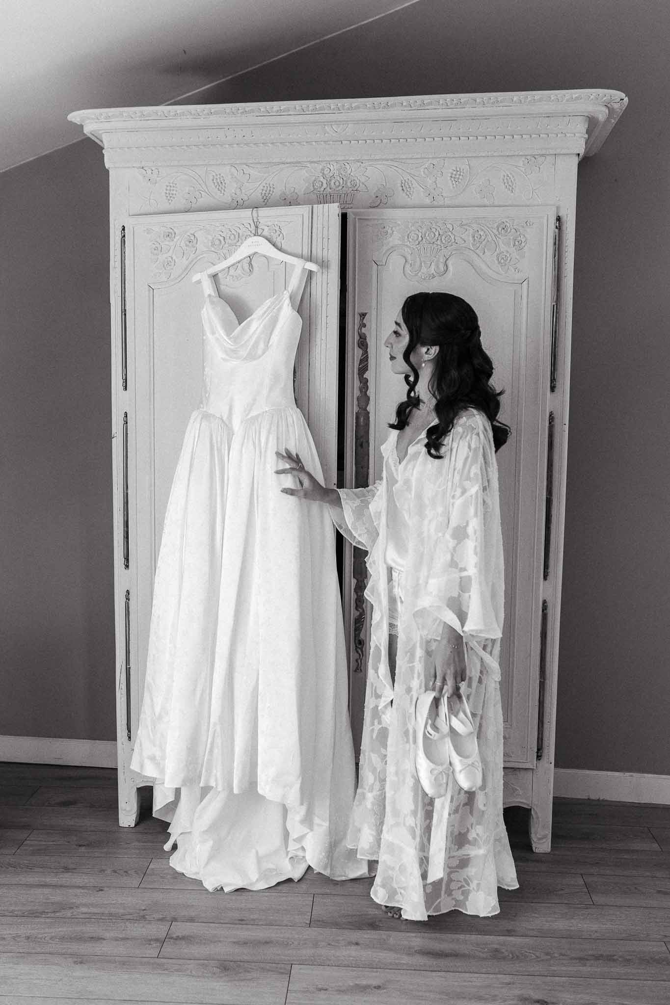 Black and white of bride in lace robe touching cowl-neck gown hanging on carved white armoire door with flat shoes