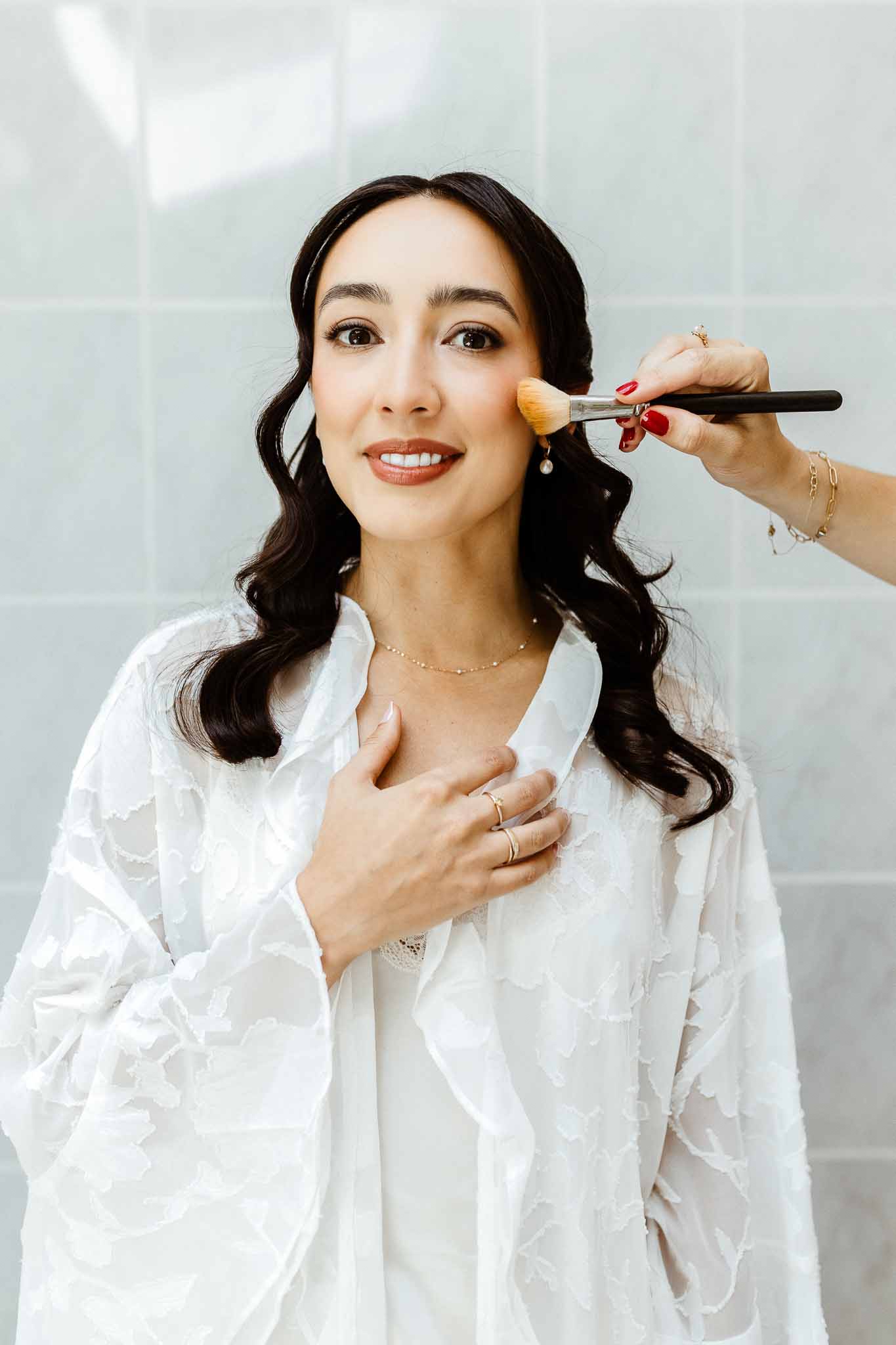 Makeup artist applies powder to smiling bride in white lace robe with gold necklace and pearl earrings