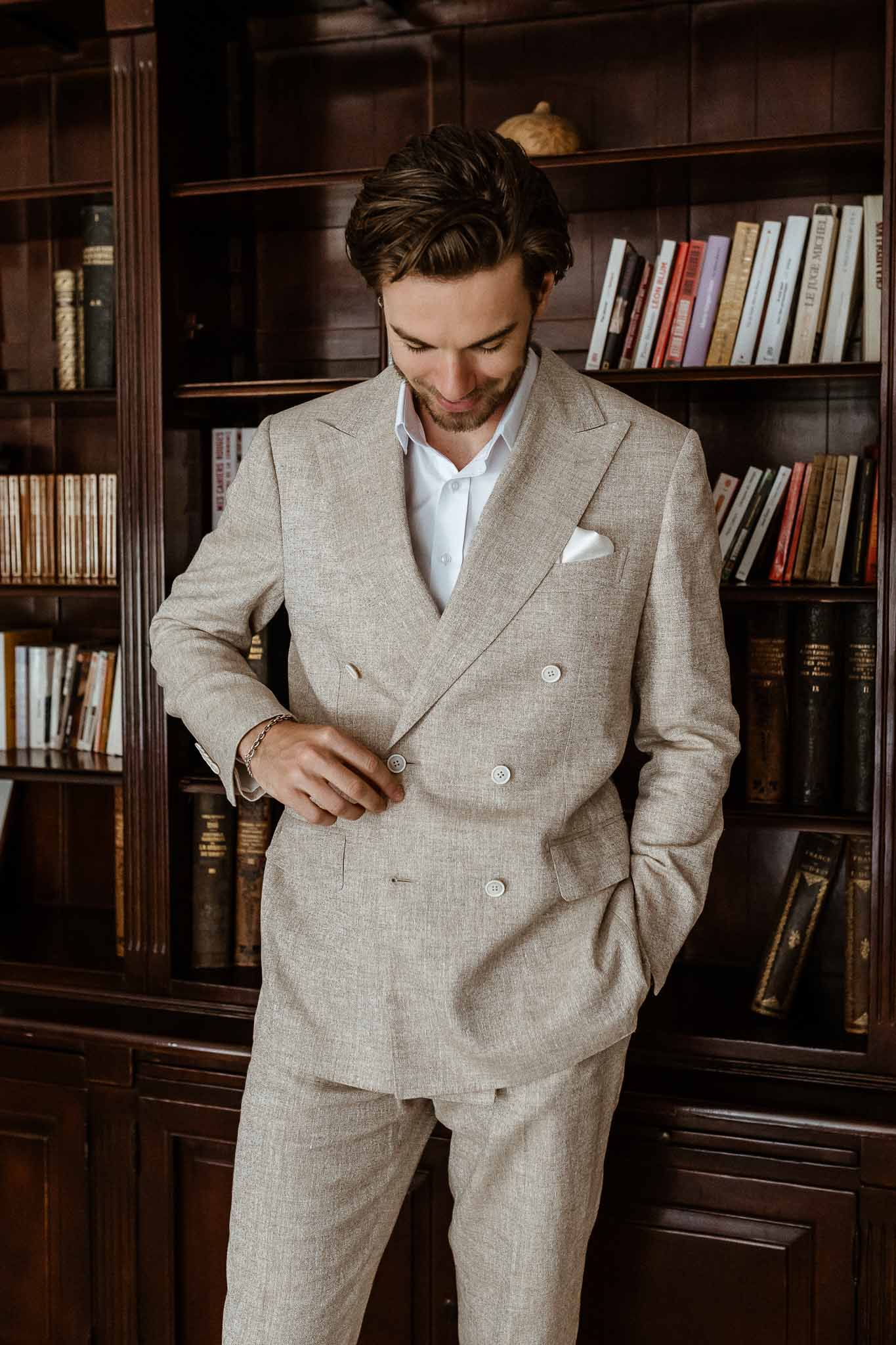 Groom buttoning beige linen double-breasted suit in wood-paneled library with floor-to-ceiling bookshelves