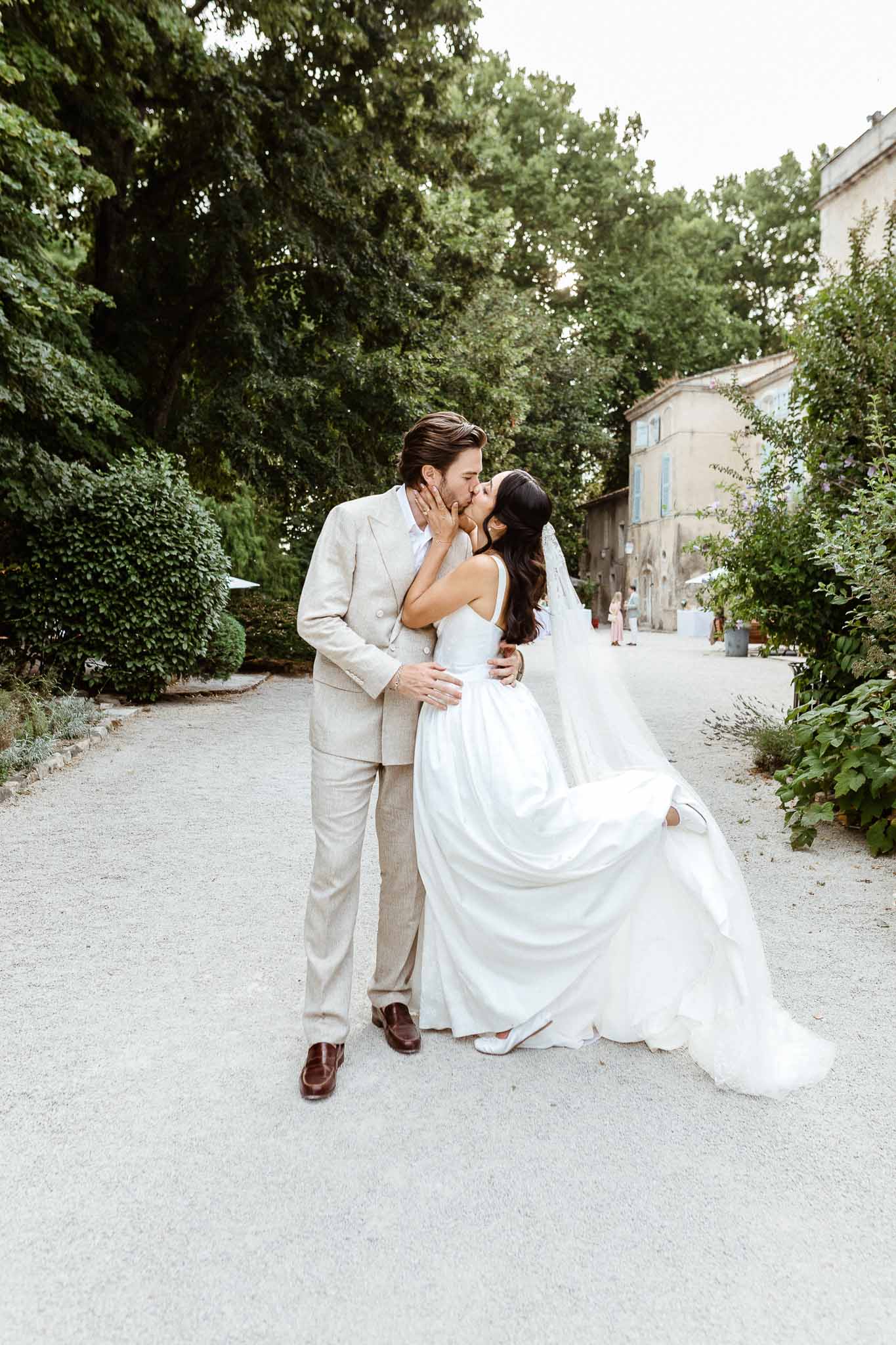 Bride in white ballgown and groom in sand linen suit kissing on gravel pathway at Provencal property