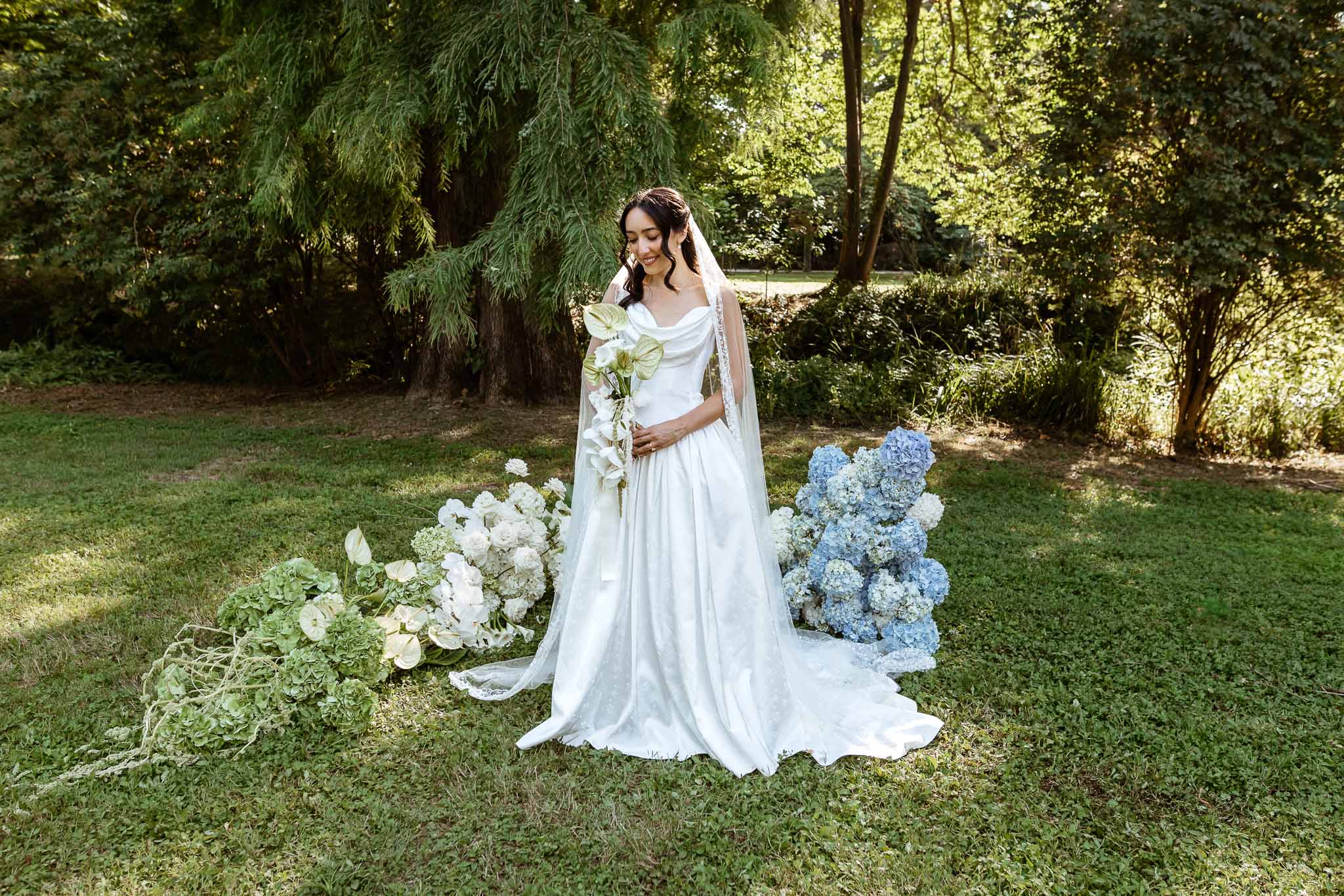 Bride in off-the-shoulder ball gown with cathedral veil holding white orchid bouquet surrounded by ground-level florals