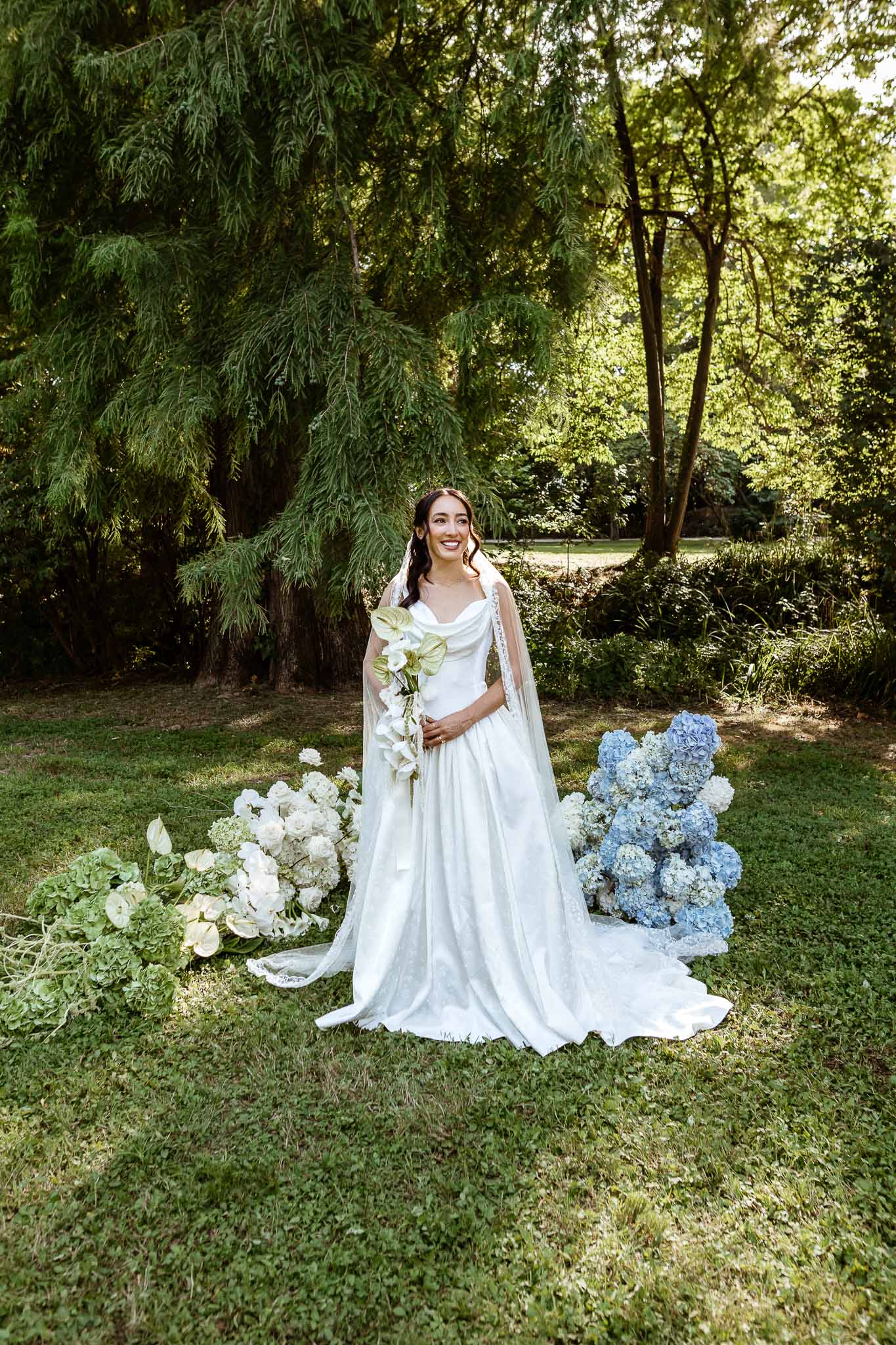 Bride in A-line gown with cape veil and calla lily bouquet beside white-to-blue gradient floral display