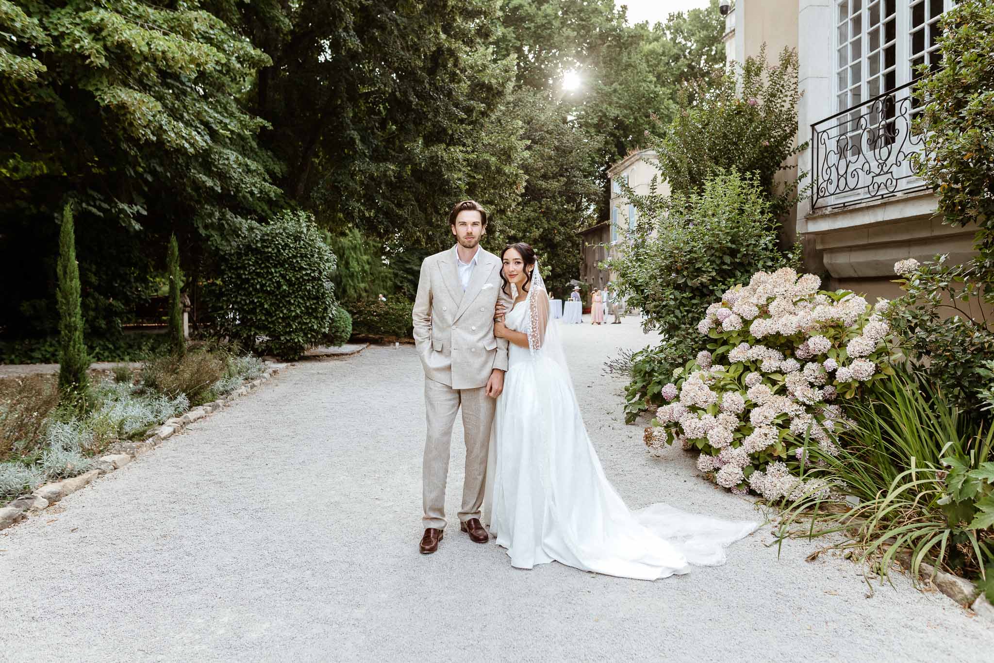 Bride in off-shoulder gown with cathedral veil and groom in beige linen suit on chateau gravel path