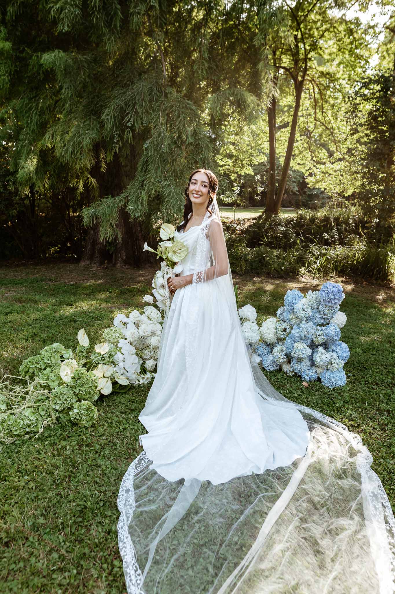 Bride in A-line lace gown with cathedral veil holding white anthurium bouquet beside blue hydrangea installation