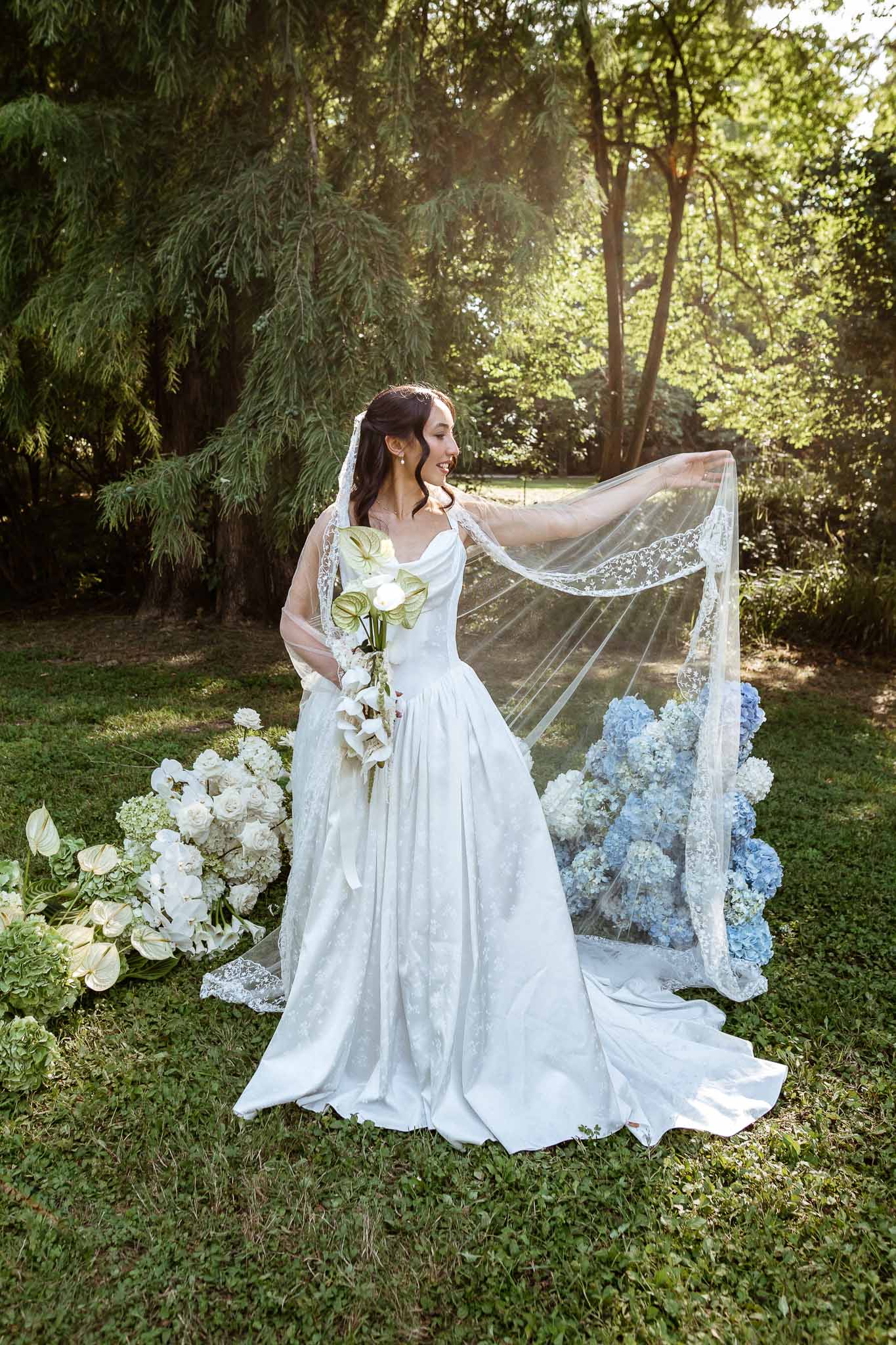 Bride lifting cathedral veil with white anthurium bouquet framed by hydrangea ground arrangements in garden