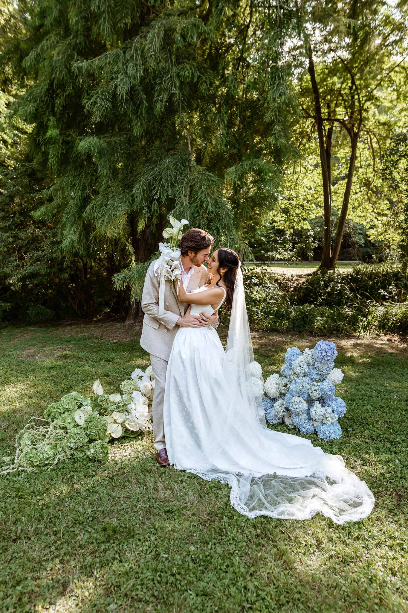 Couple nose to nose with blue and white hydrangea ground arrangements, bride in lace-edged cathedral veil