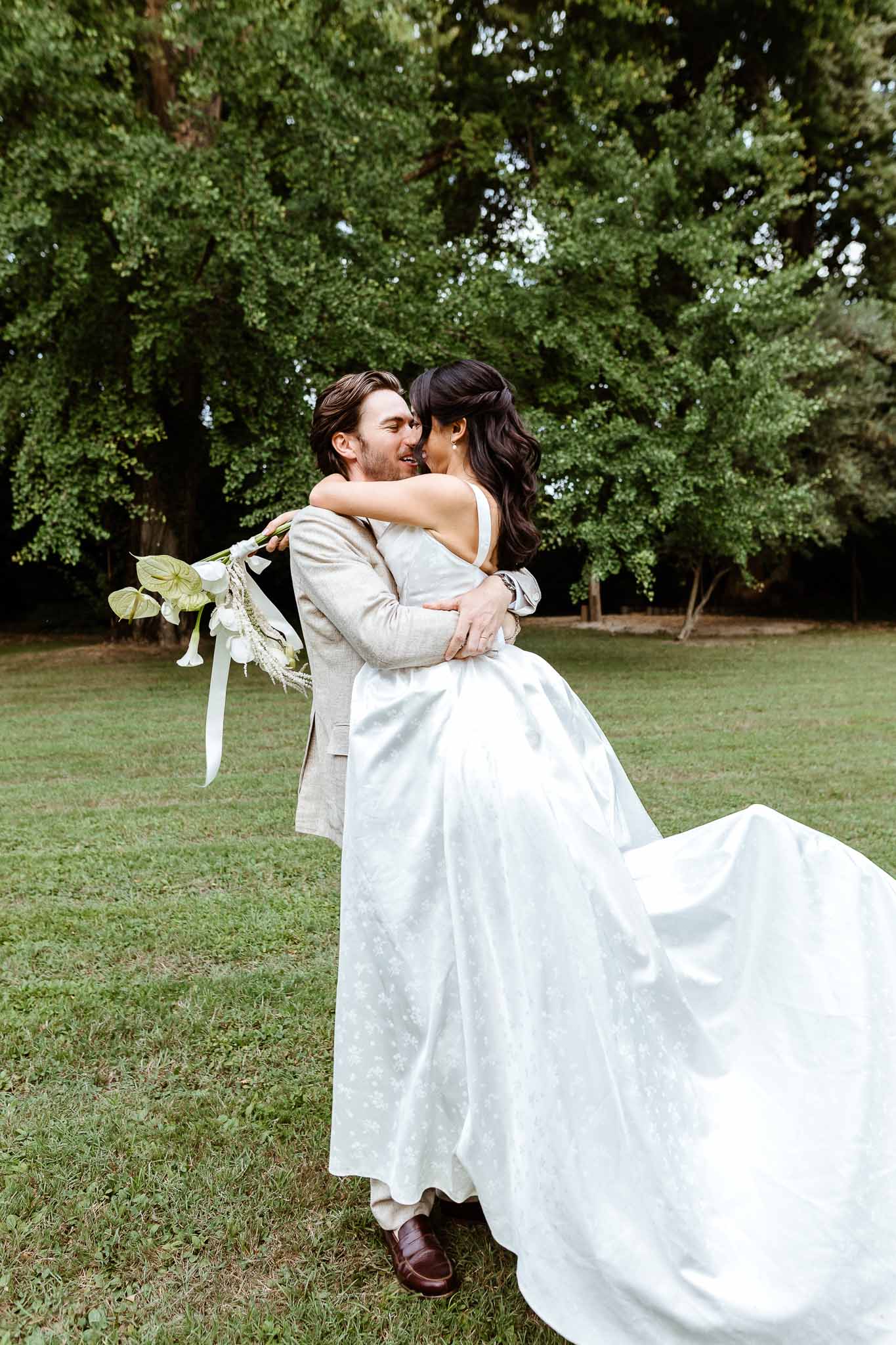 Groom lifting bride on lawn, bride in jacquard ballgown holding white calla lily bouquet with ribbons