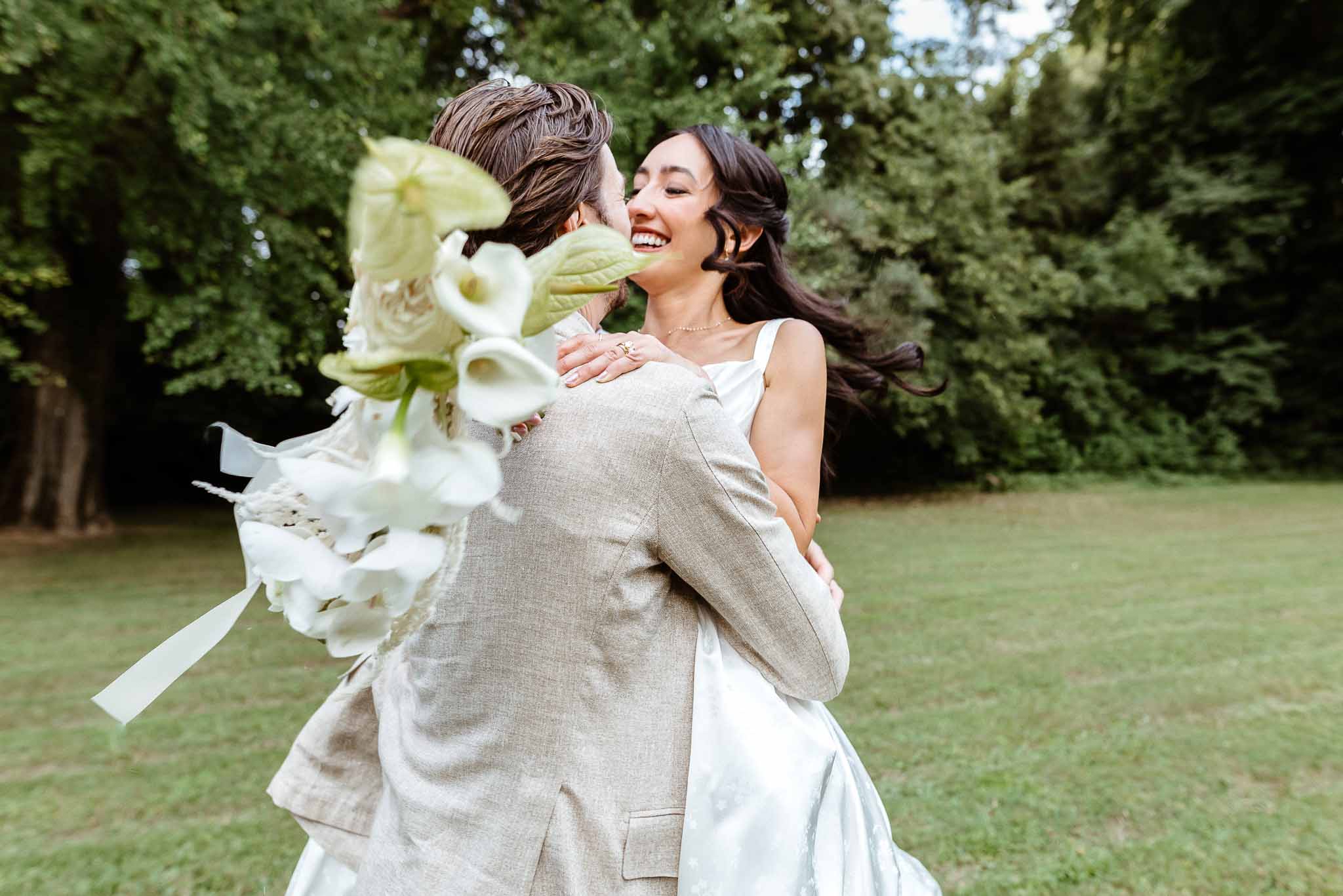 Groom lifting laughing bride in white satin gown with calla lily bouquet blurred in foreground
