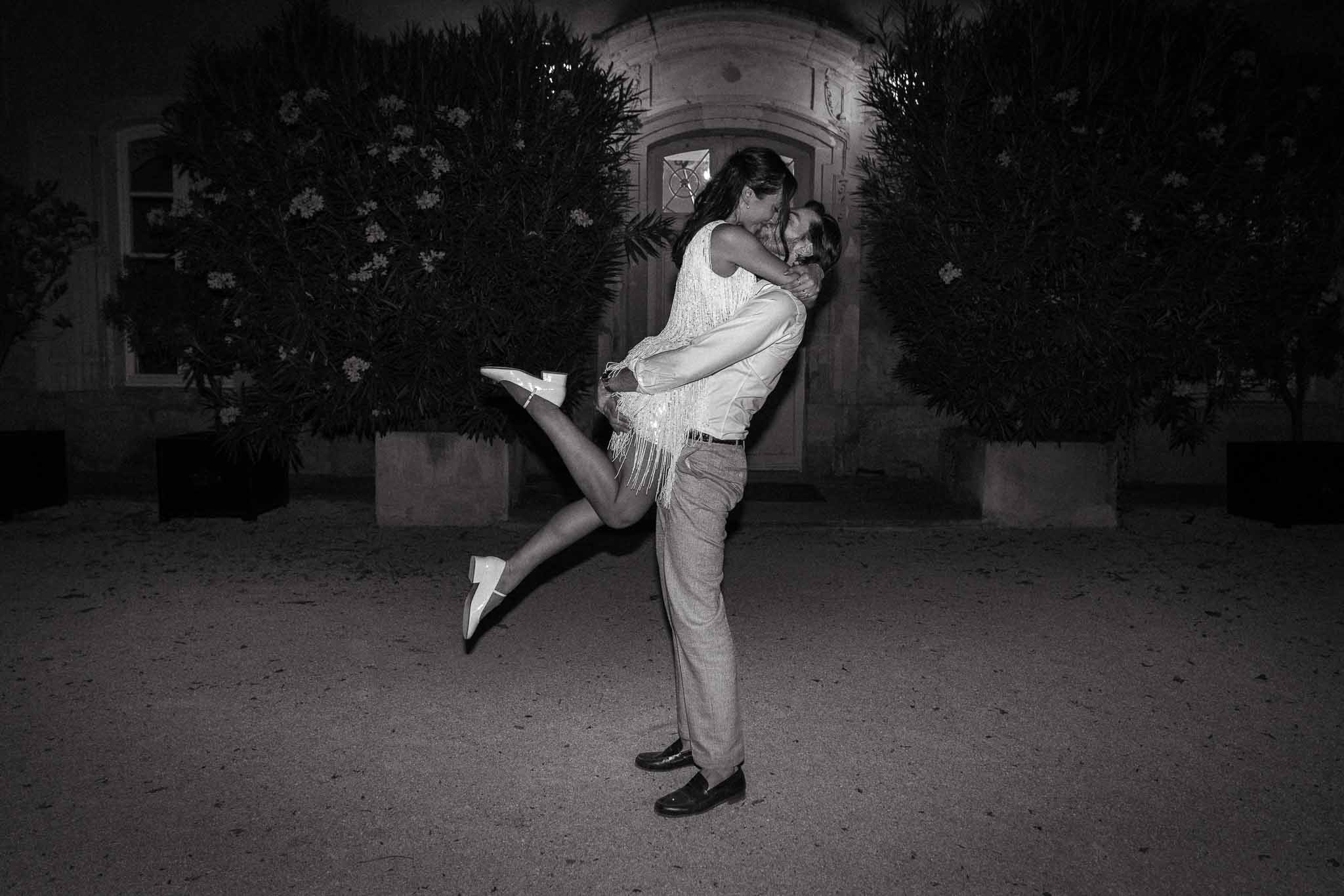 Black and white nighttime portrait of groom lifting bride in fringed mini dress before lit chateau doorway