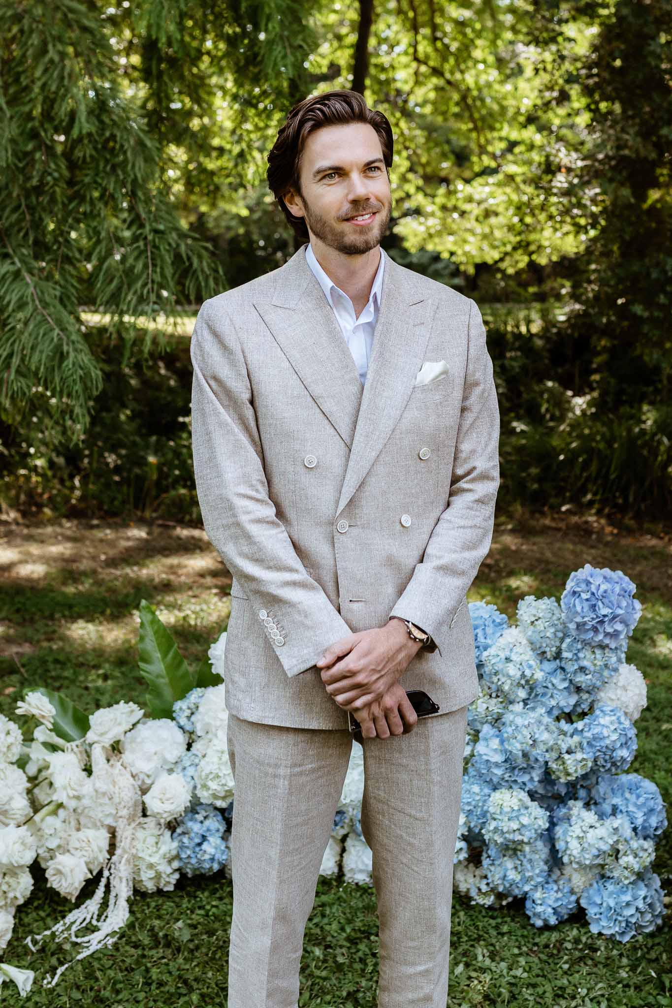 Groom in sand linen suit standing before blue and white hydrangea arrangement at outdoor ceremony