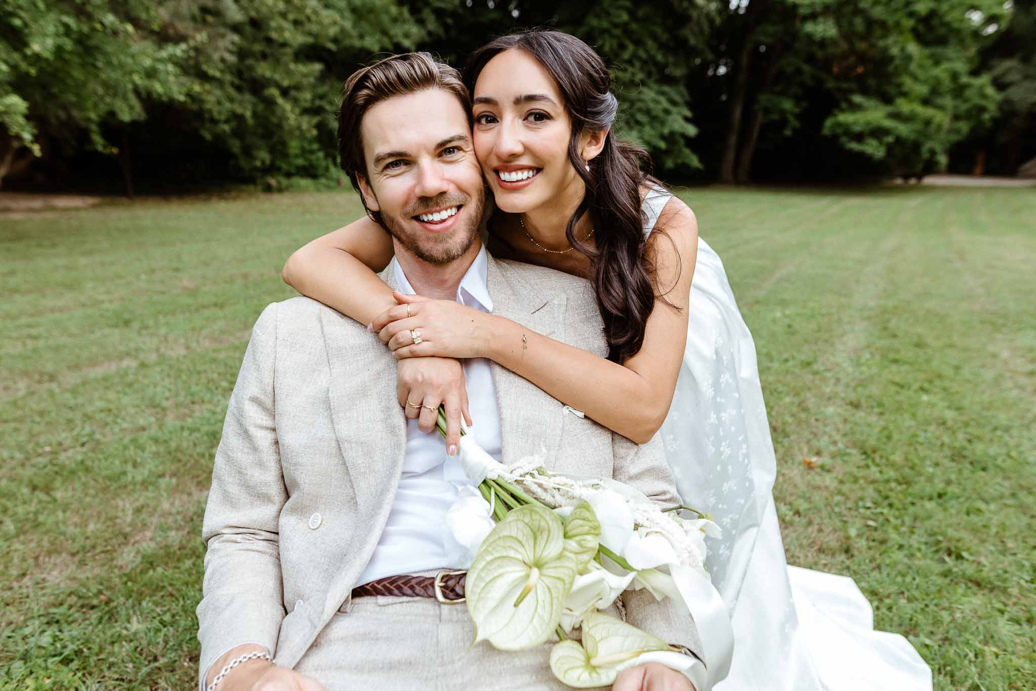 Couple seated on grass with bride hugging groom from behind holding white calla lily and green anthurium bouquet