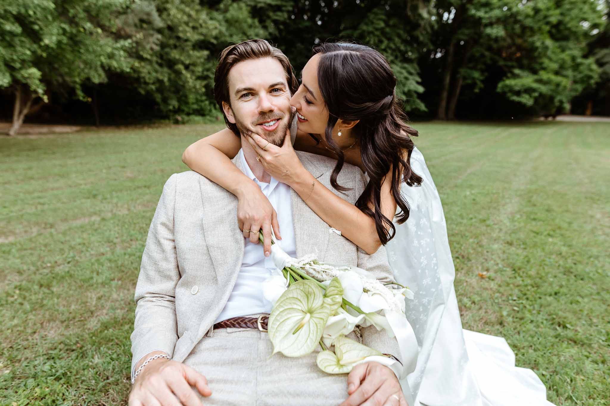 Bride whispering to seated groom on garden lawn, holding white calla lily and anthurium bouquet