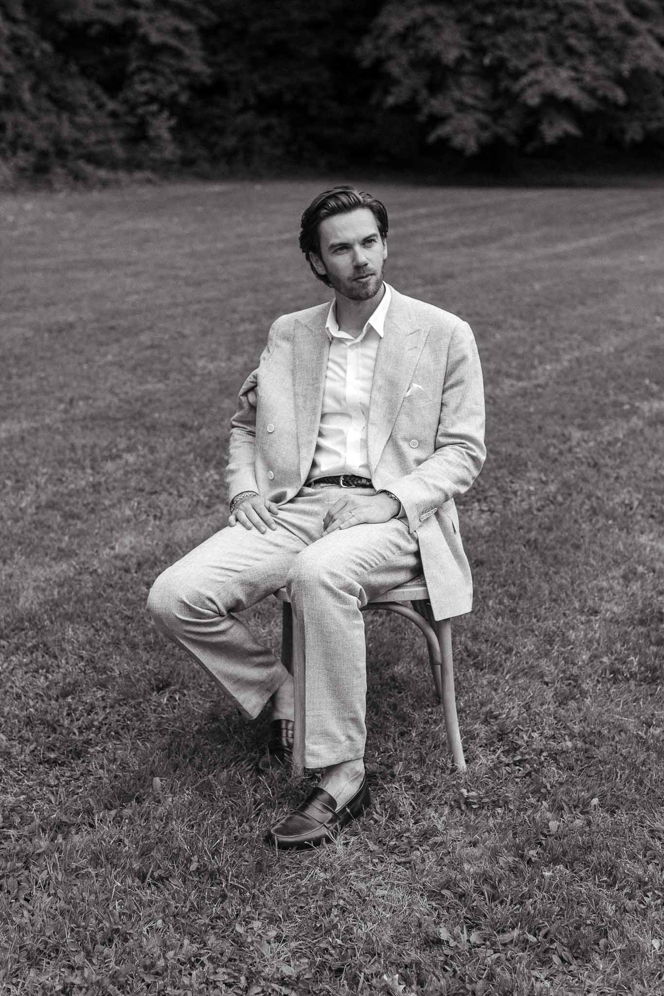 Black and white full-length portrait of groom in double-breasted suit seated on bistro chair on lawn