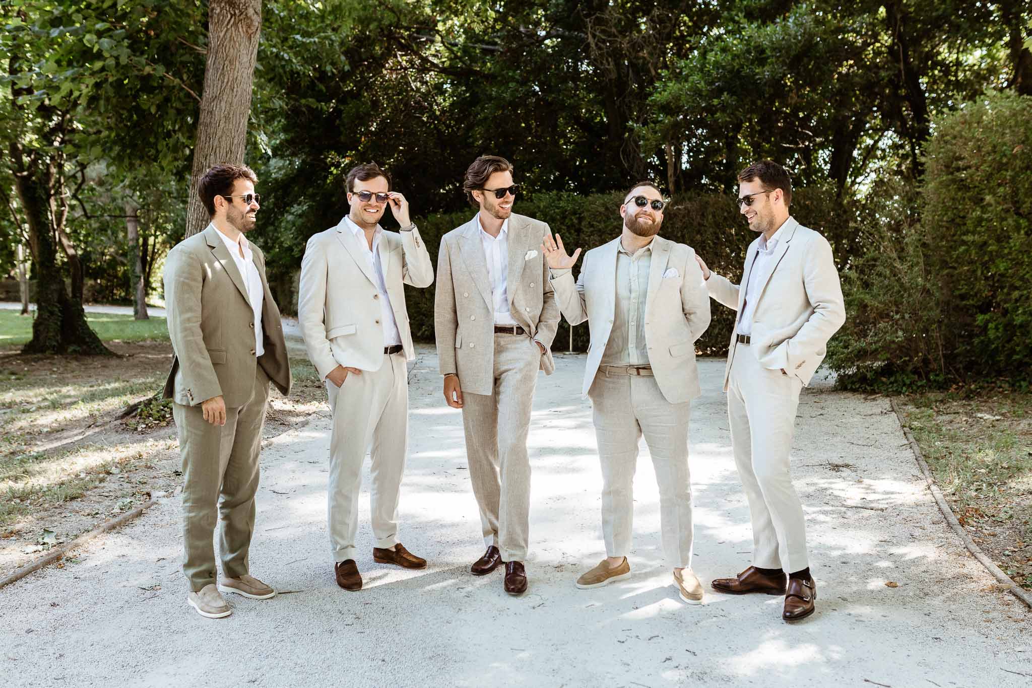 Groom in cream suit with four groomsmen in beige linen suits laughing on gravel path