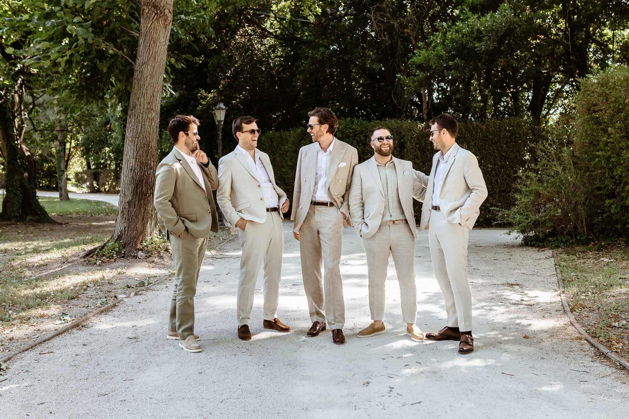 Five groomsmen in beige linen suits laughing together on gravel path in park grounds