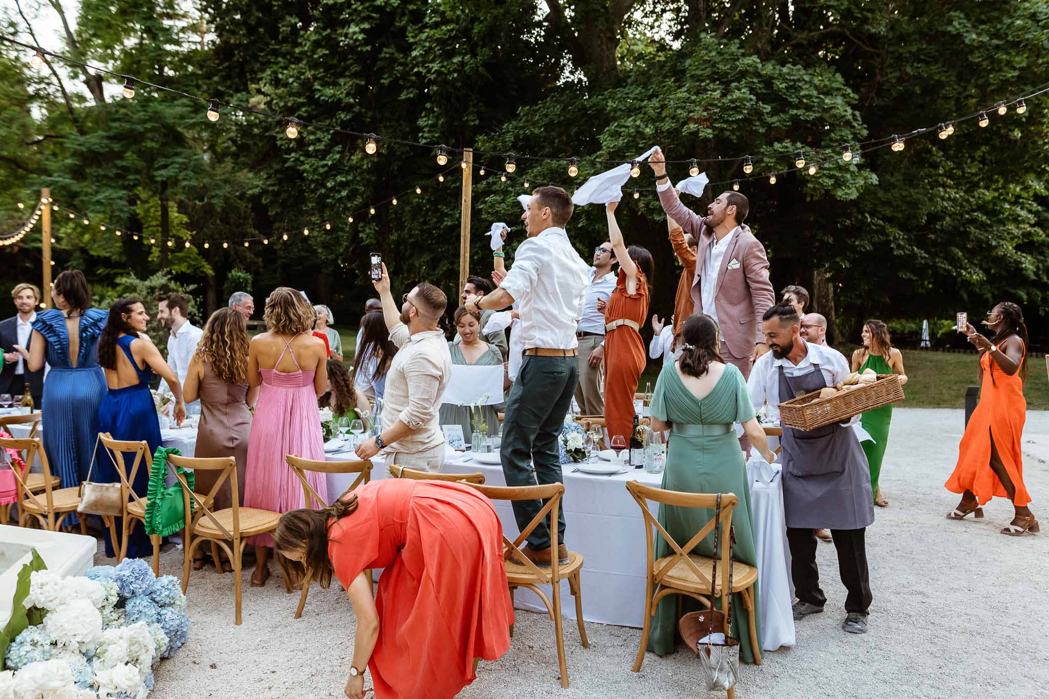 Guests waving white napkins at dusk reception with blue hydrangea centerpieces and string lights