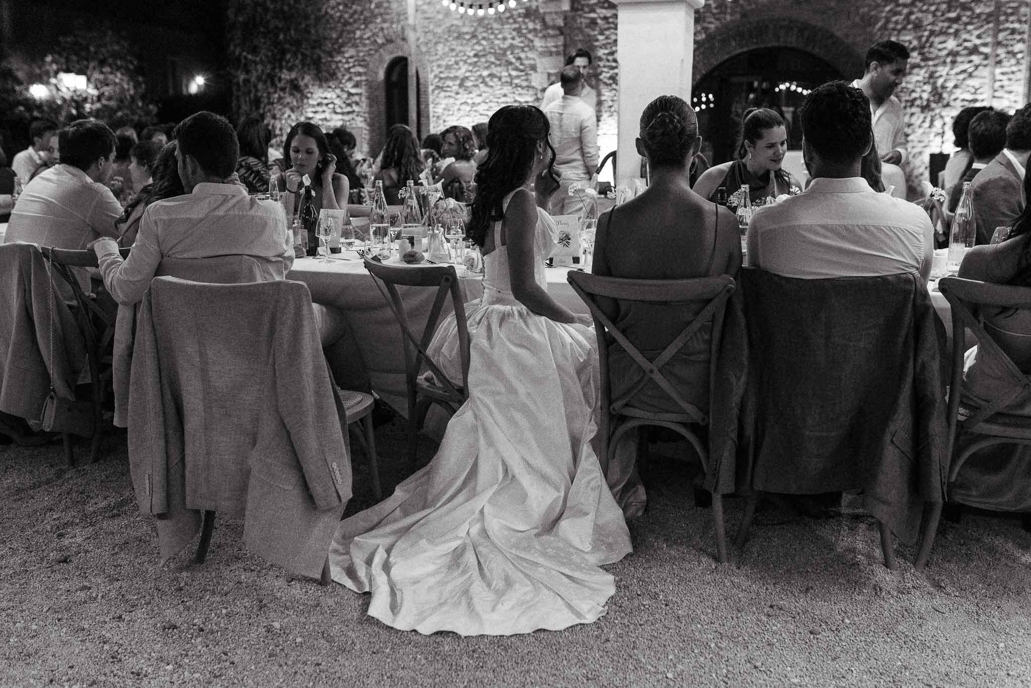 Bride seated among fifteen guests at round tables in stone courtyard lit by string lights at night in B&W
