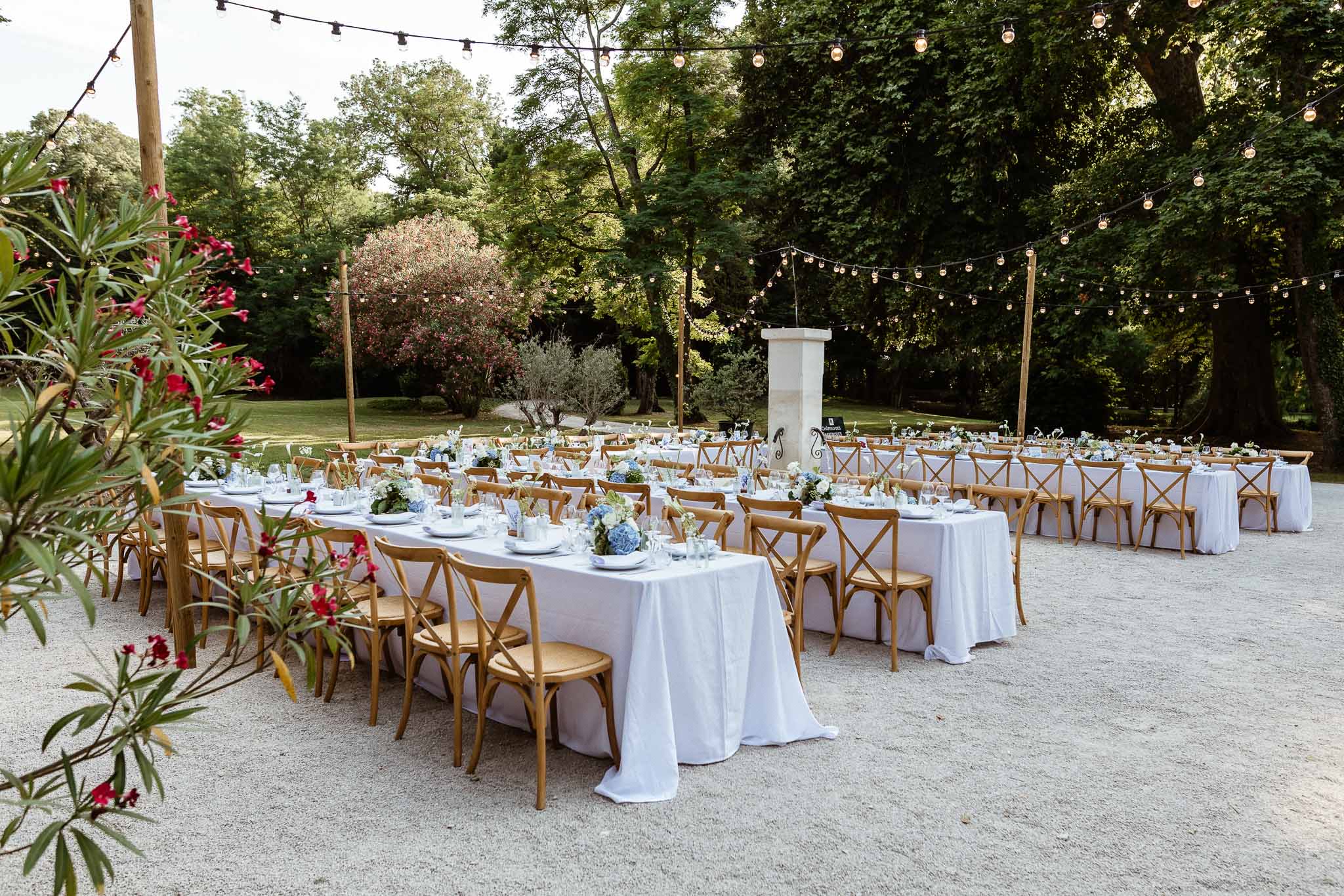 Long banquet tables on gravel terrace with blue hydrangea centerpieces and Edison bulb string lights overhead