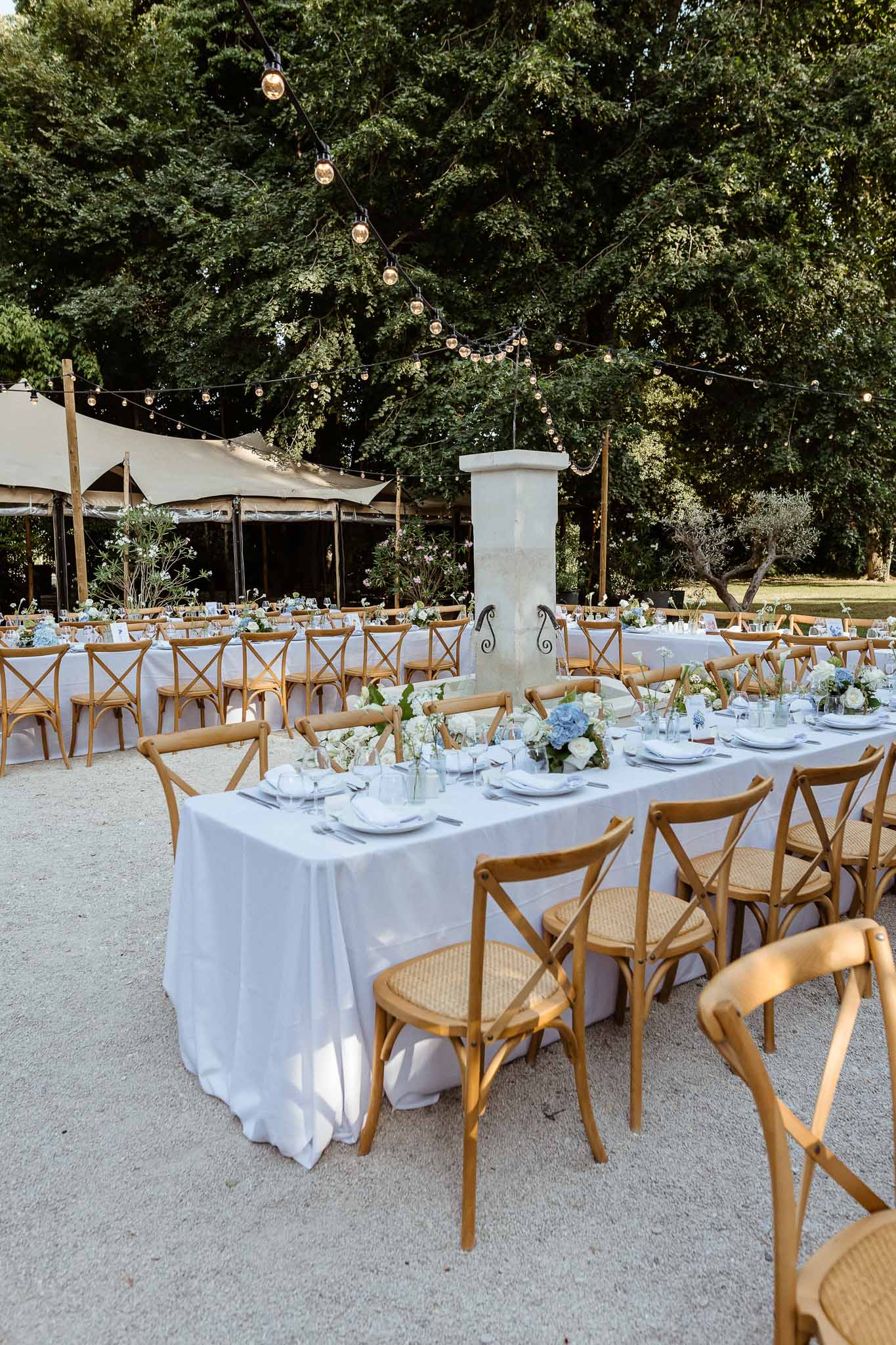 Outdoor reception with blue-grey linen tables, blue hydrangea centerpieces, and festoon lights on gravel
