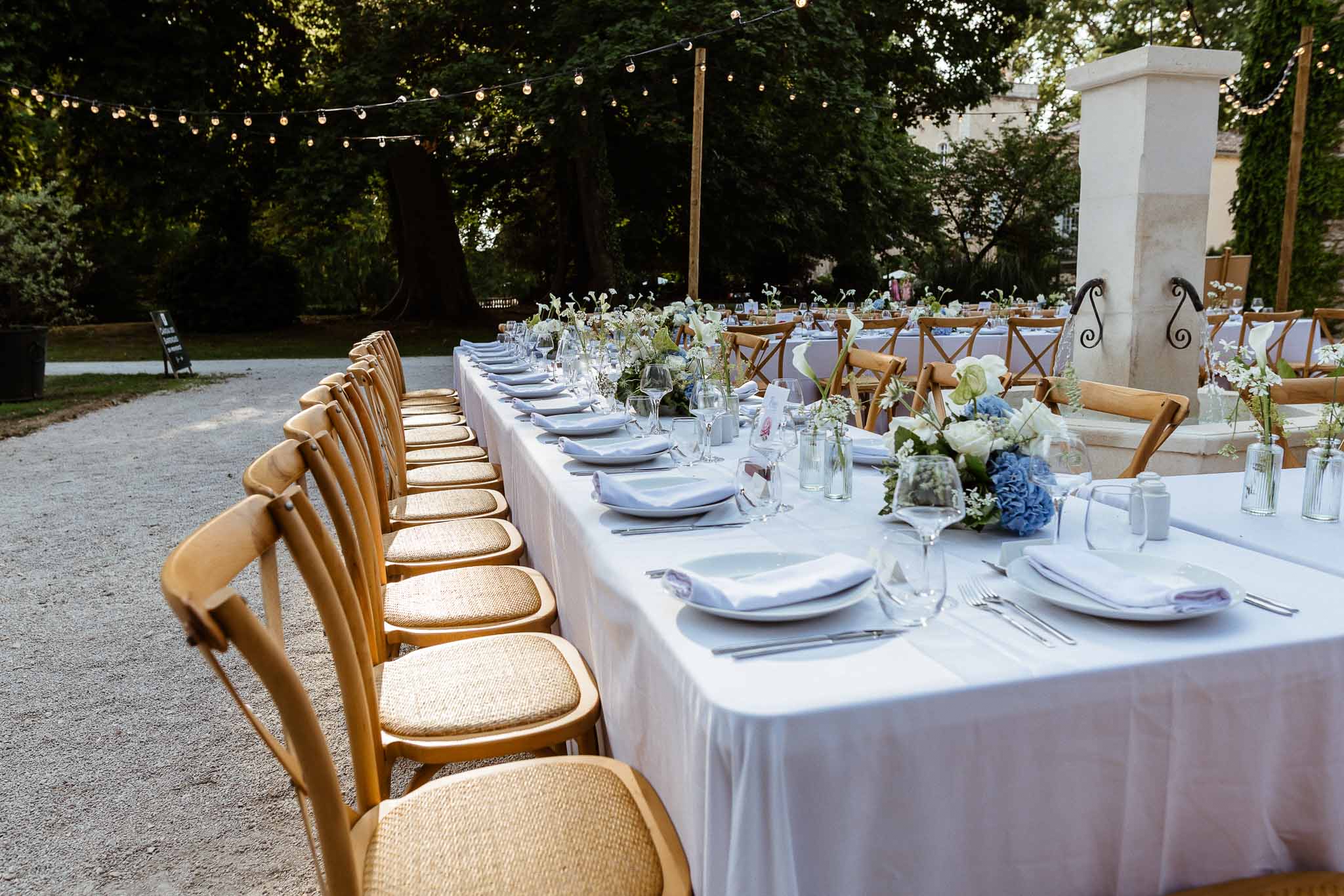 Long banquet table with blue hydrangea centerpieces cross-back chairs and string lights at dusk