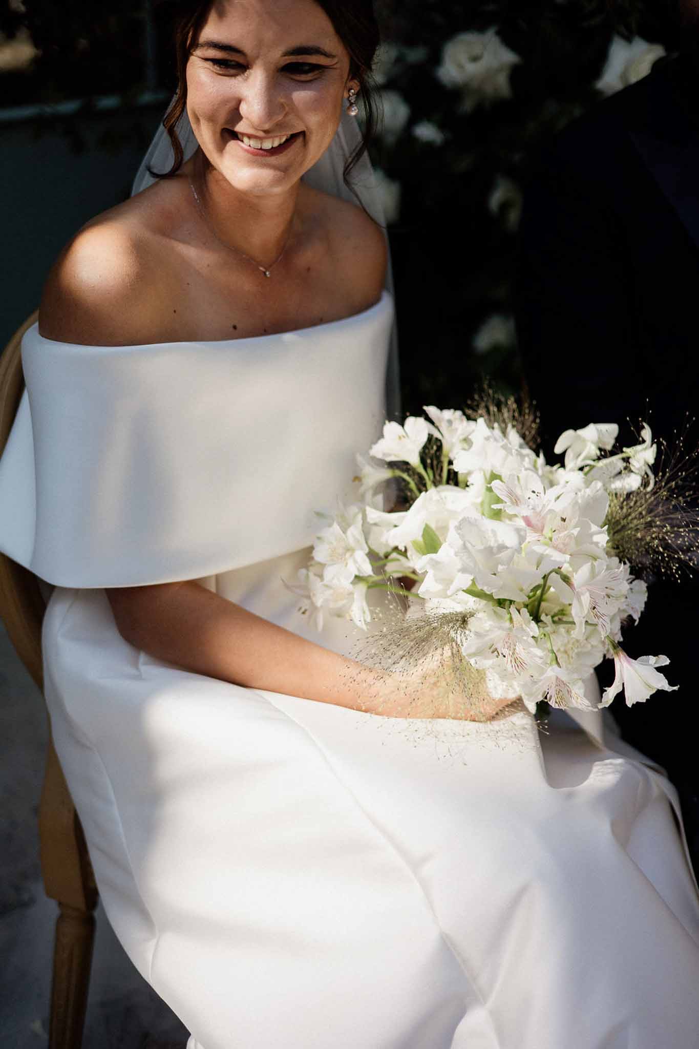 Laughing bride in off-shoulder white gown holding bouquet of white alstroemeria and dried grasses outdoors