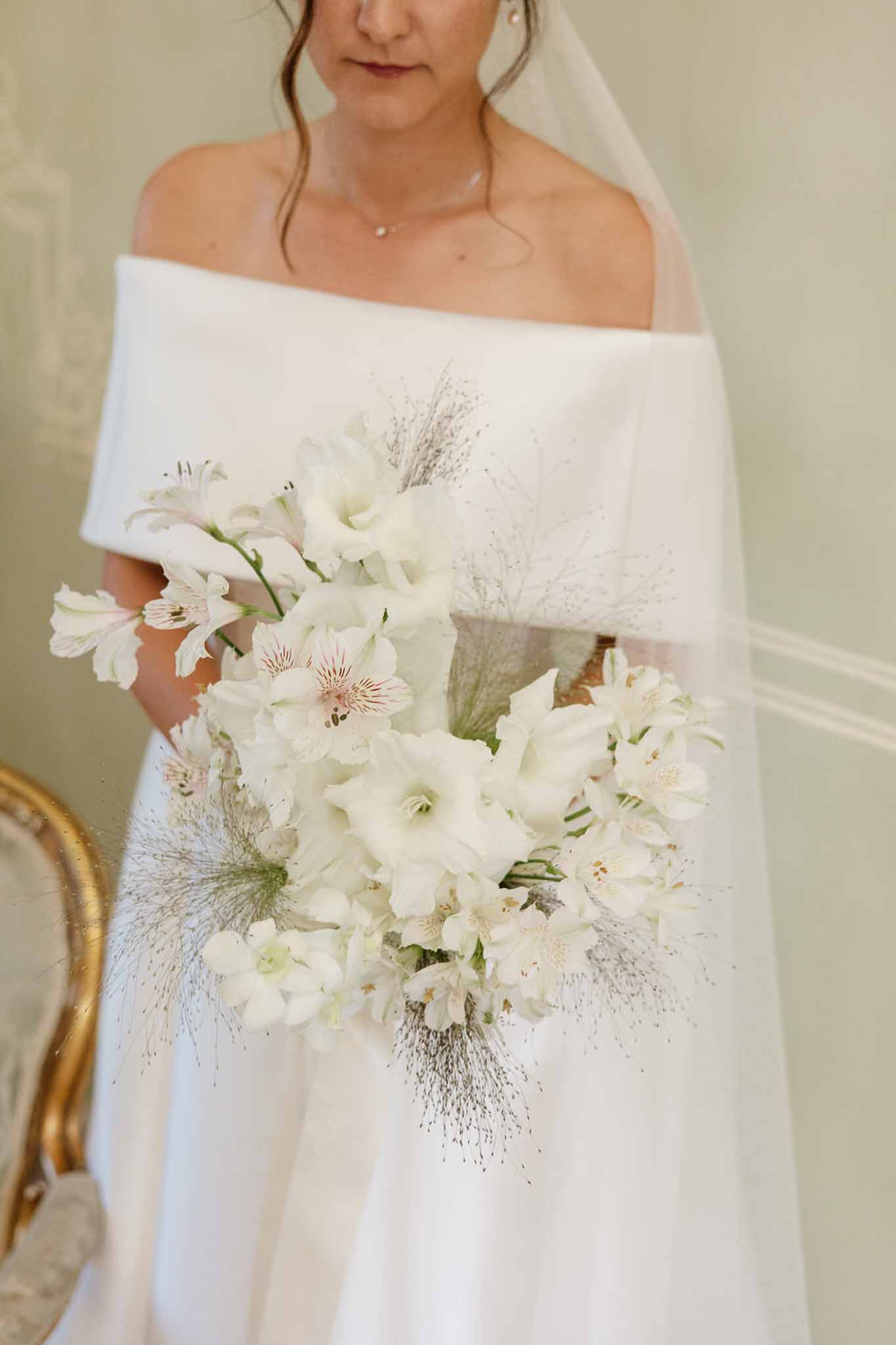 Bride holding all-white bouquet of gladioli and alstroemeria with wispy greenery and pearl necklace