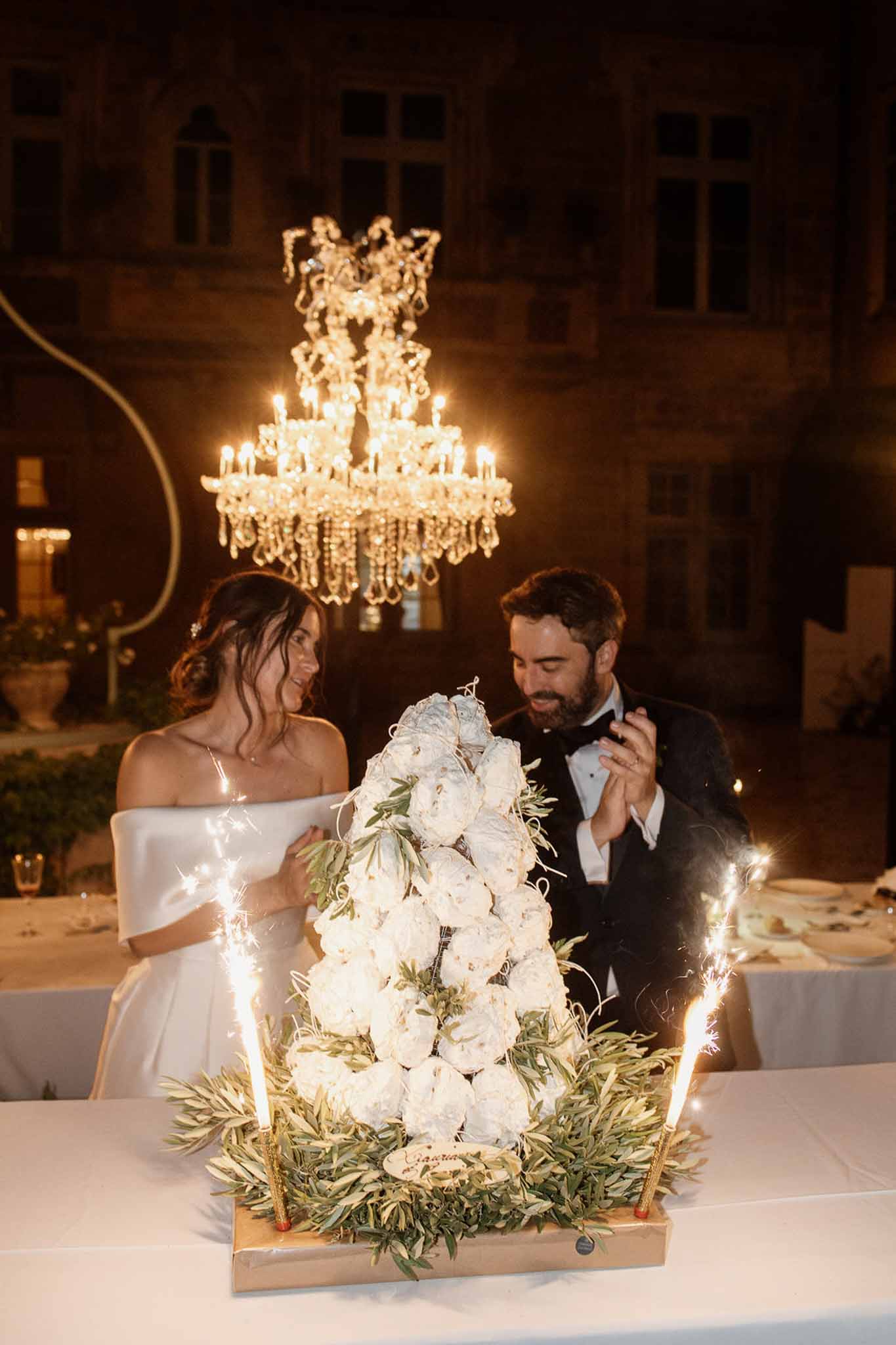 Bride and groom at sweetheart table reacting to croquembouche cake with lit sparklers under outdoor chandelier