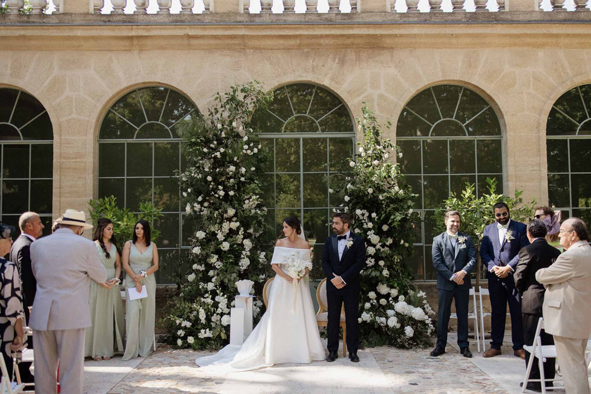 Ceremony before orangery with white rose floral columns, sage bridesmaids, and navy groomsmen on white chairs