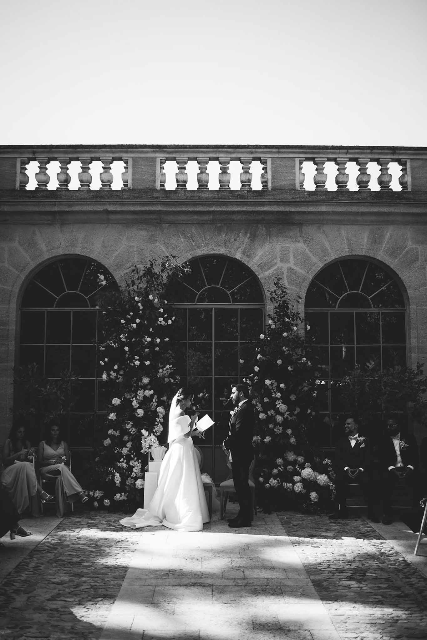 Black and white image of bride reading vows at outdoor ceremony flanked by large floral installations