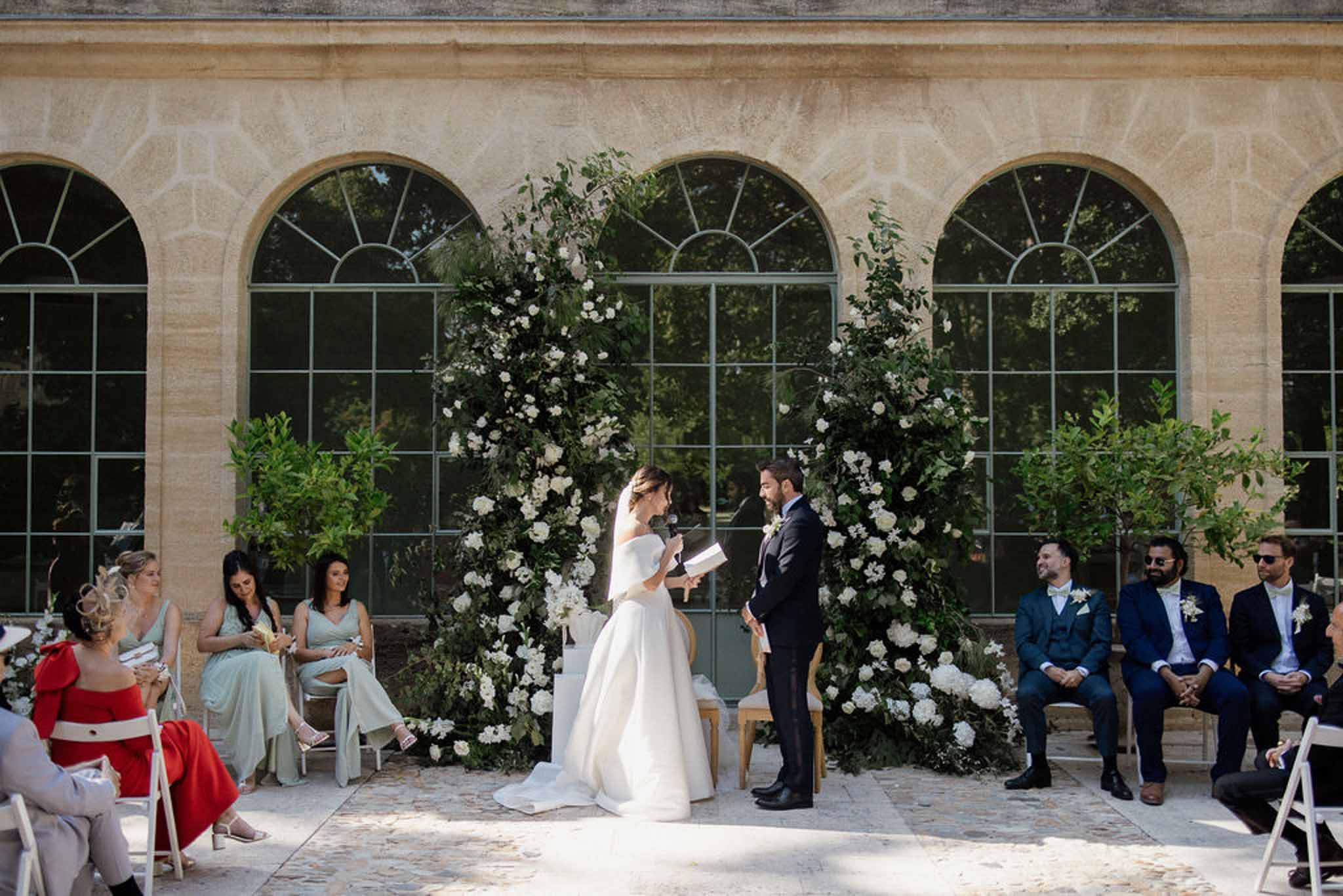 Outdoor ceremony with bride reading vows under circular white rose and greenery arch at French orangerie