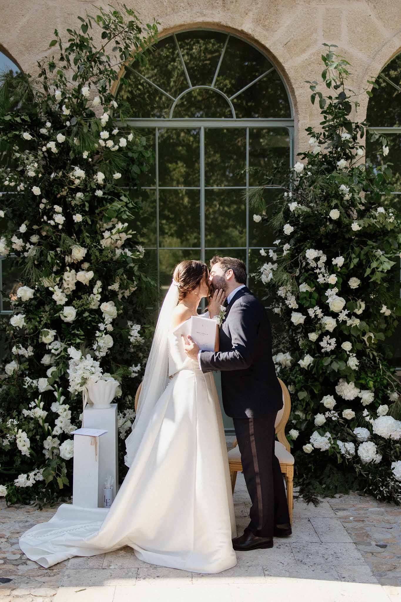 First kiss before arched stone window flanked by white rose and peony floral columns with green foliage