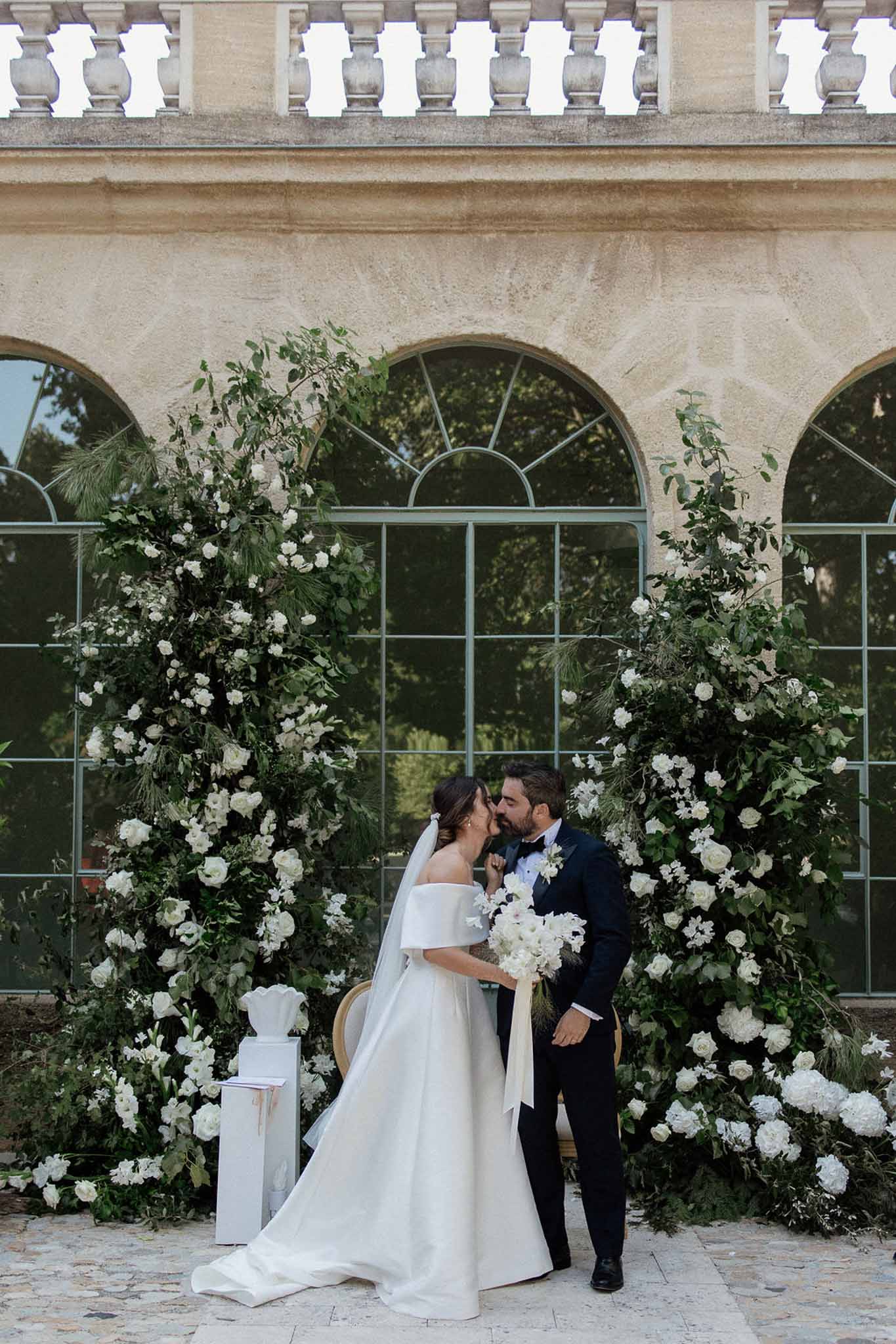First kiss under asymmetrical white rose and greenery installations before chateau orangerie arched windows