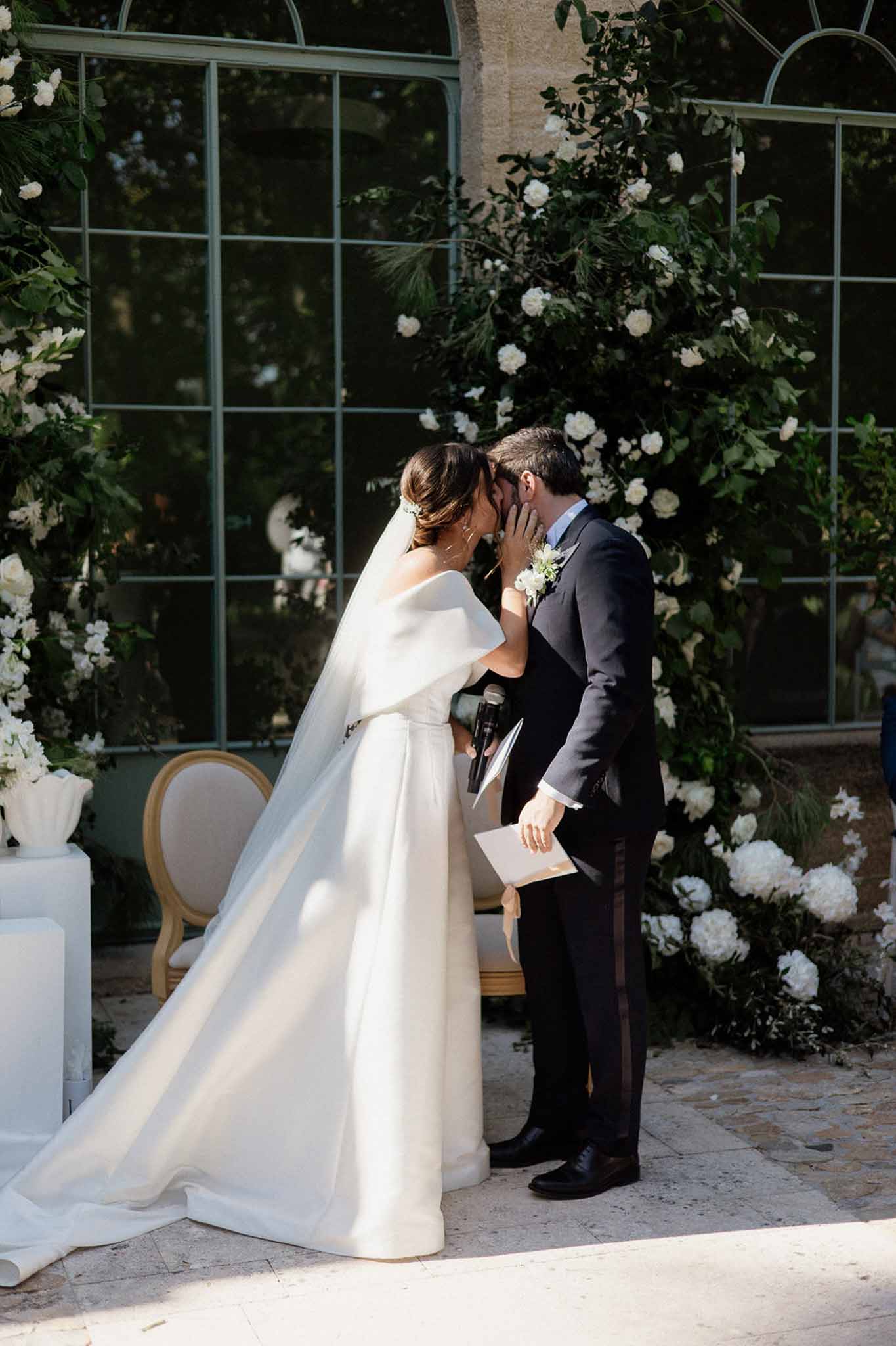 First kiss under green foliage arch with white roses before greenhouse orangery, bride in cape gown