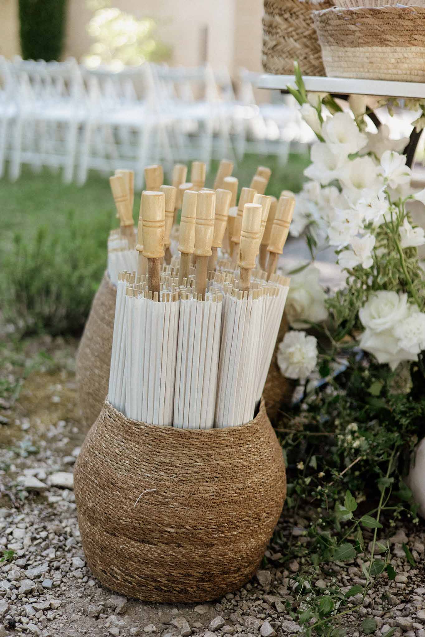 Seagrass basket of white parasols beside white rose arrangement at outdoor chateau ceremony setup