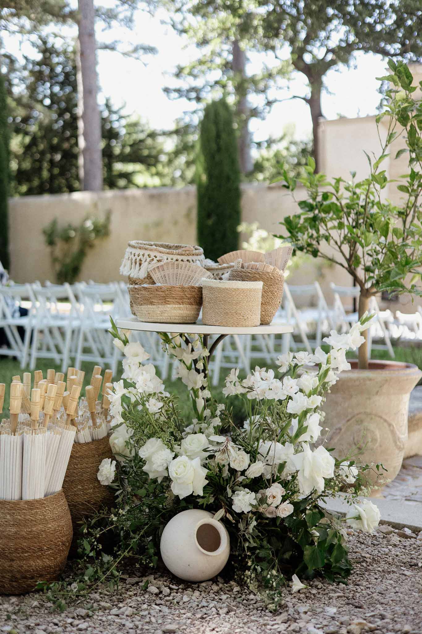 Ceremony guest station with paper fans, white parasols, and white rose arrangement on gravel