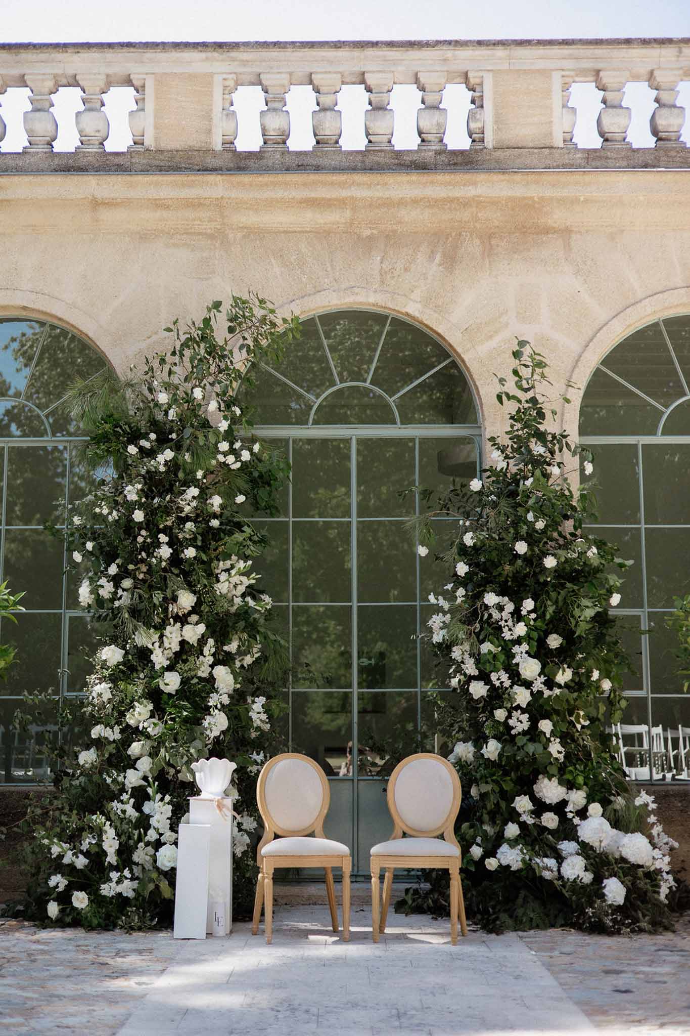 Ceremony altar with two tall green and white floral columns framing gold Louis XVI chairs before a chateau orangery