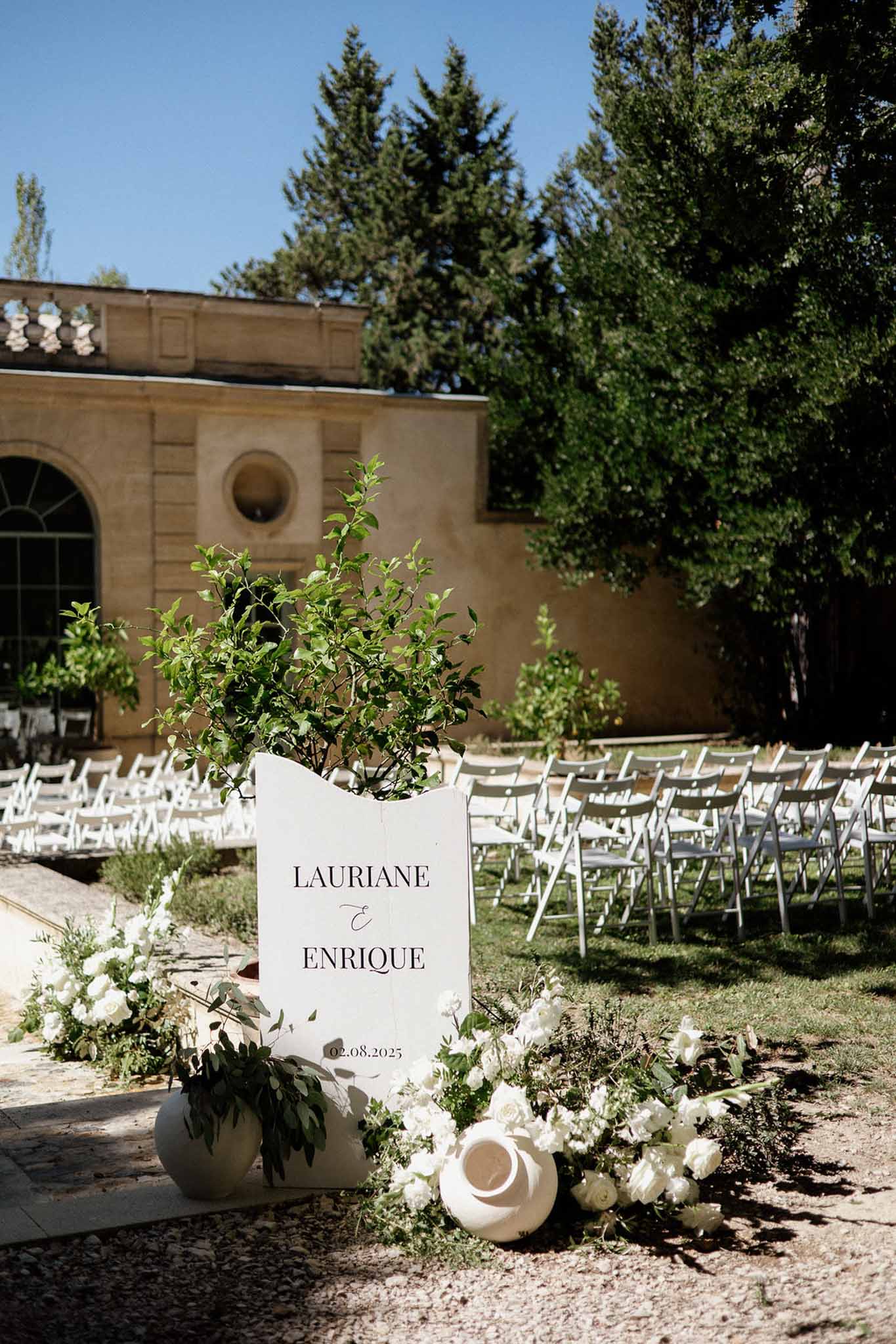 White welcome sign with rose arrangements and folding chairs on lawn before stone building with arches