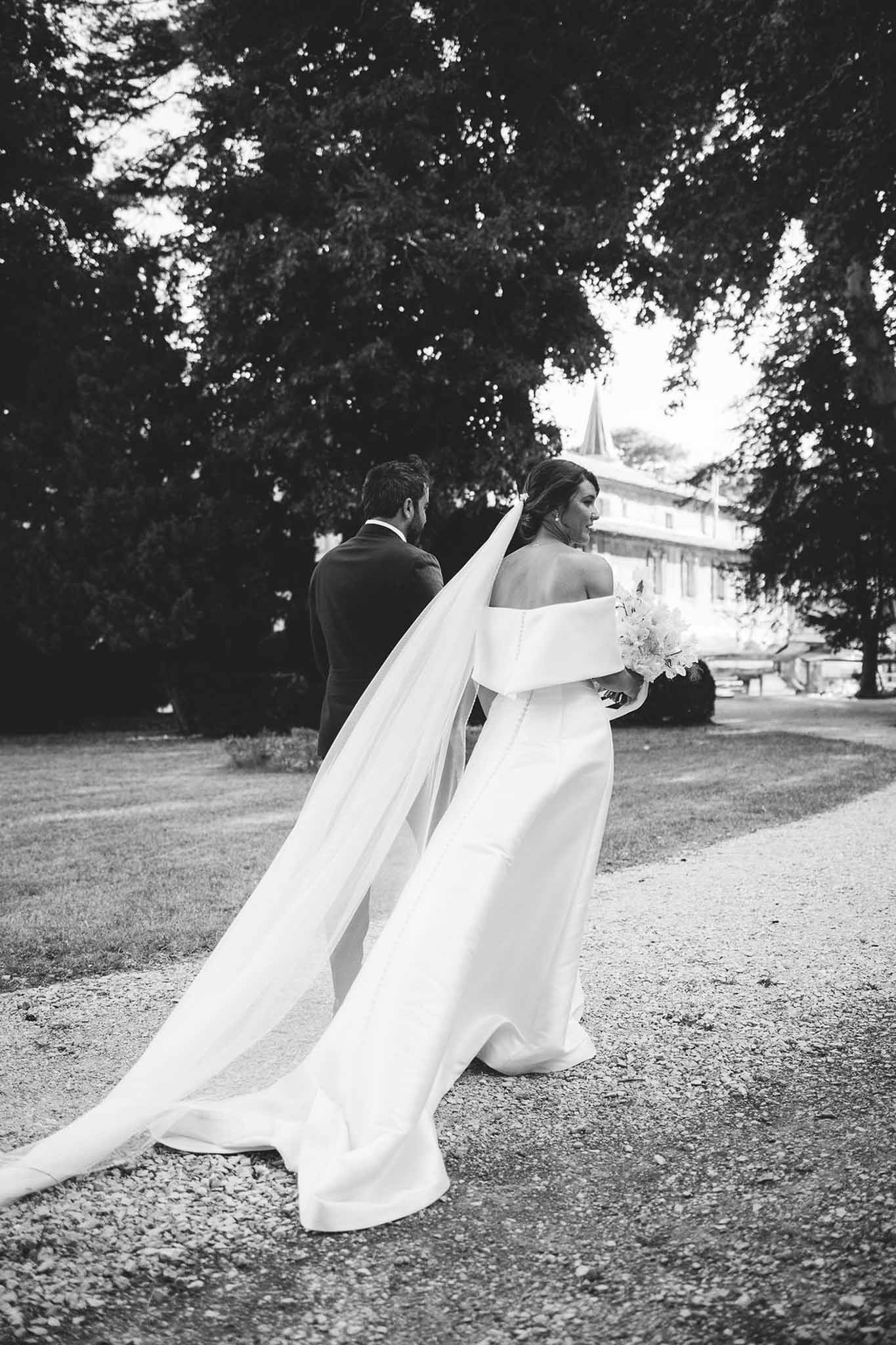 Black and white from behind of couple walking on gravel path with cathedral train and veil trailing across ground
