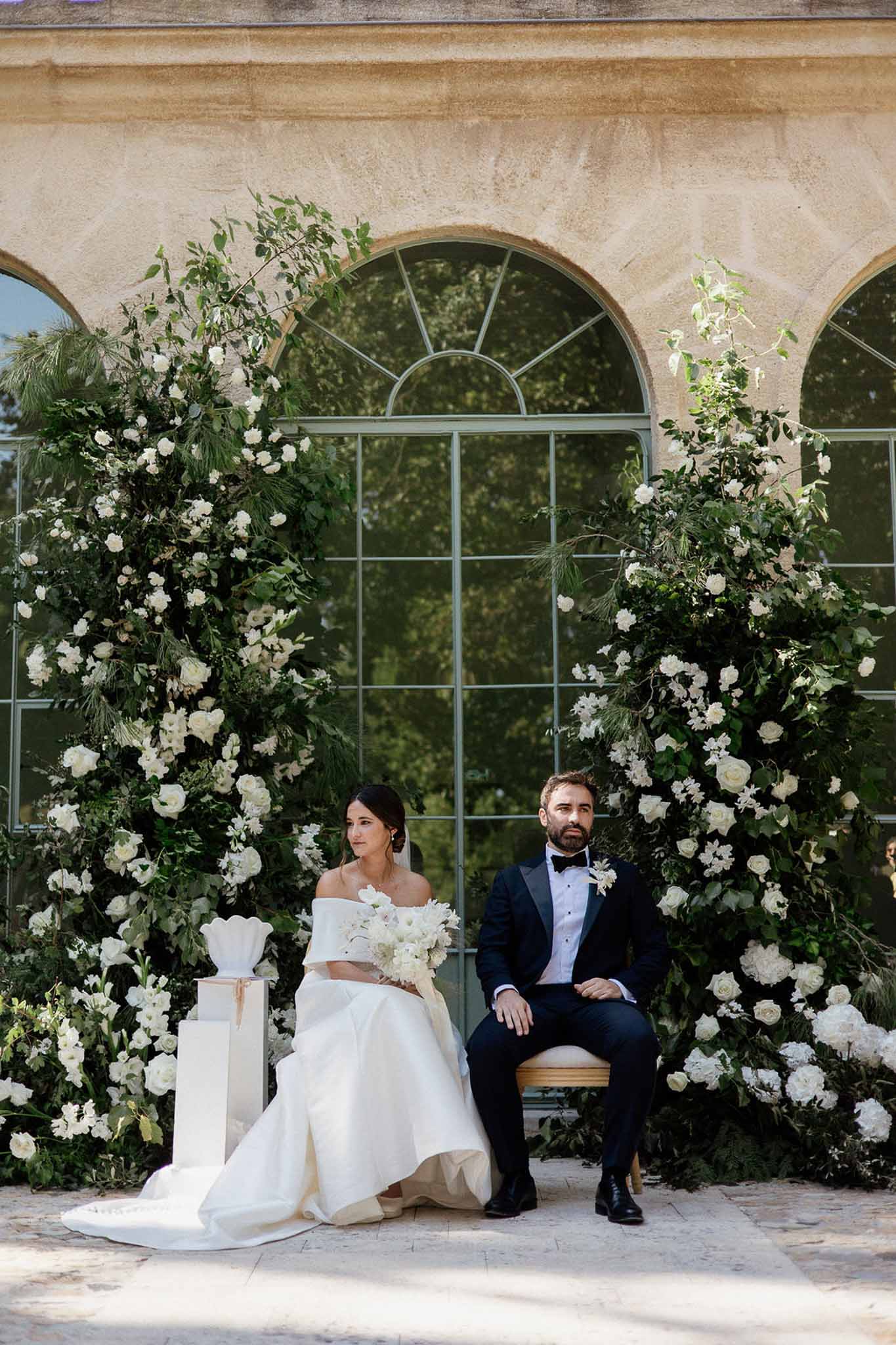 Couple seated before orangerie with tall asymmetric white rose and greenery floral columns