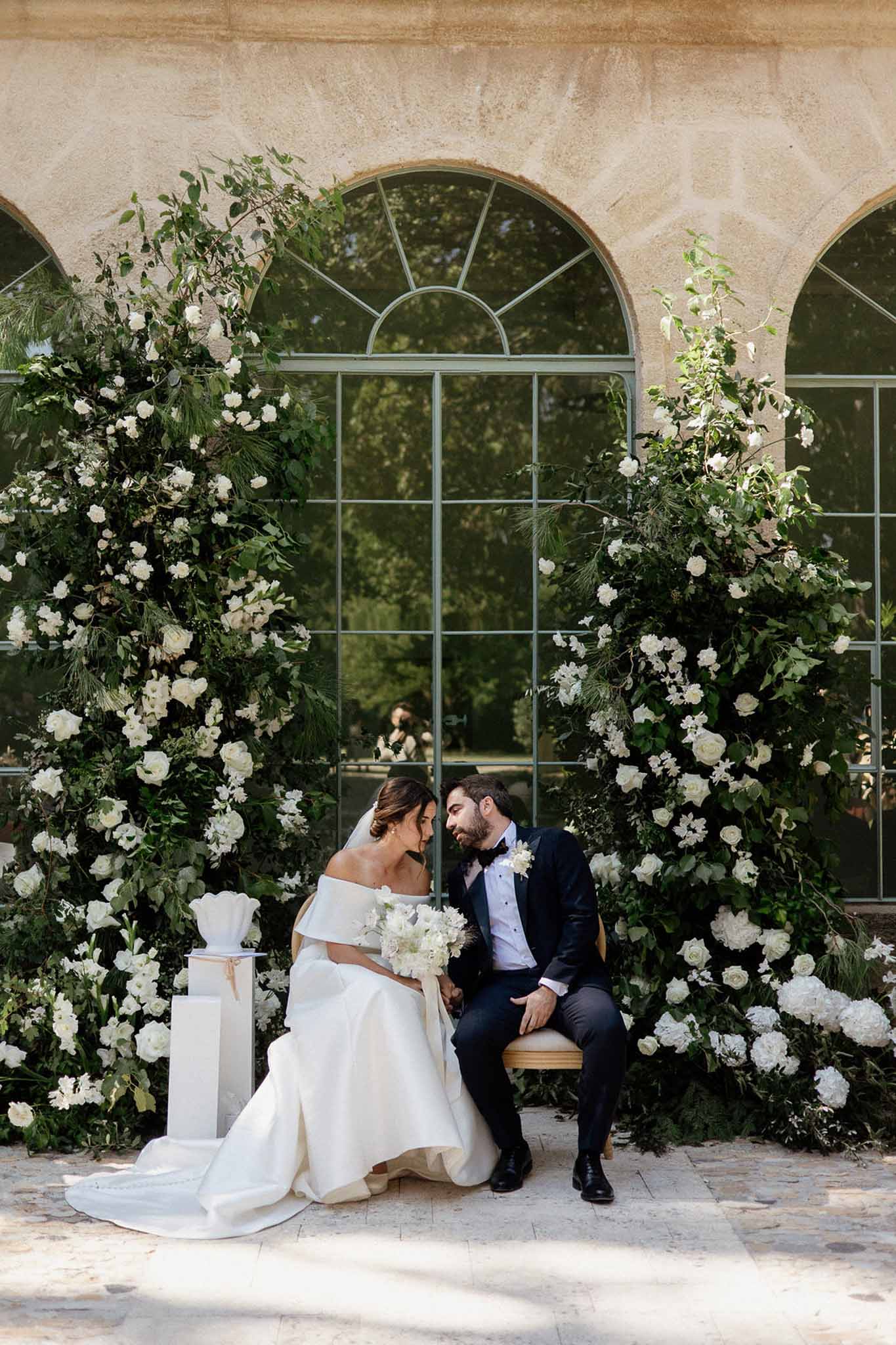 Bride and groom seated together framed by tall white and green floral installations at a chateau orangerie