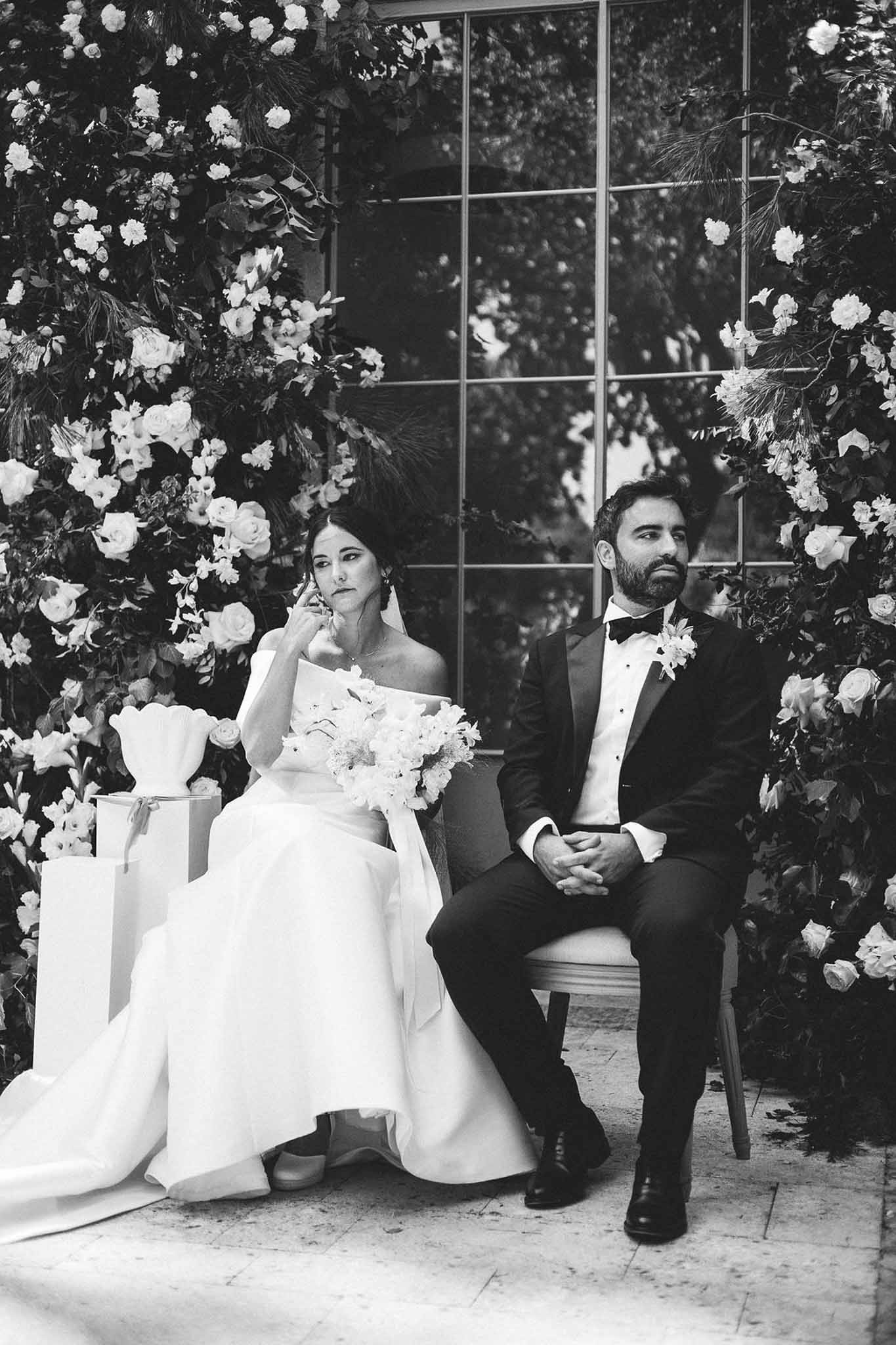 Black and white portrait of emotional bride and groom seated between dense rose floral columns