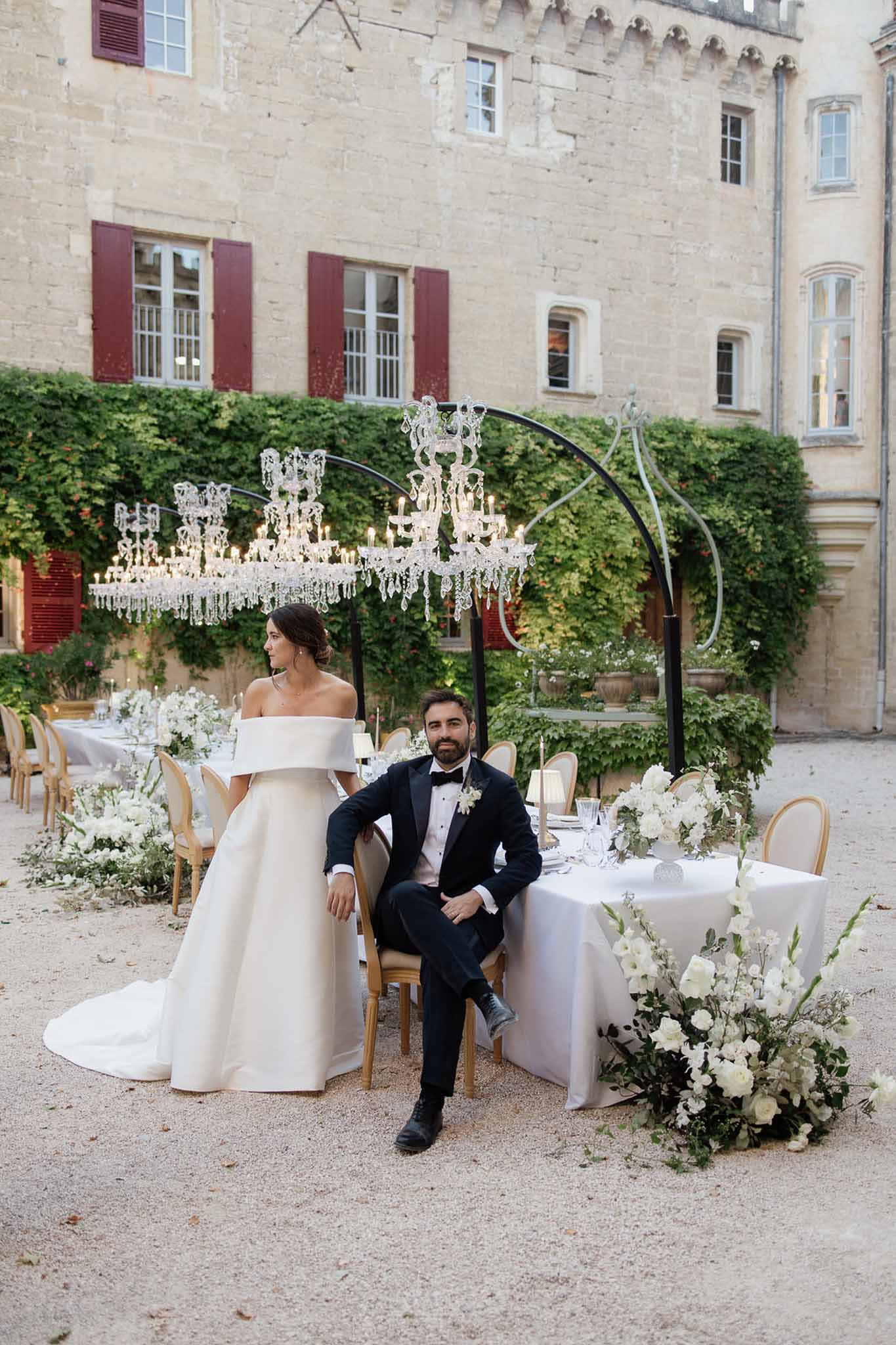 Couple at sweetheart table in chateau courtyard with crystal chandeliers white roses and gold chairs