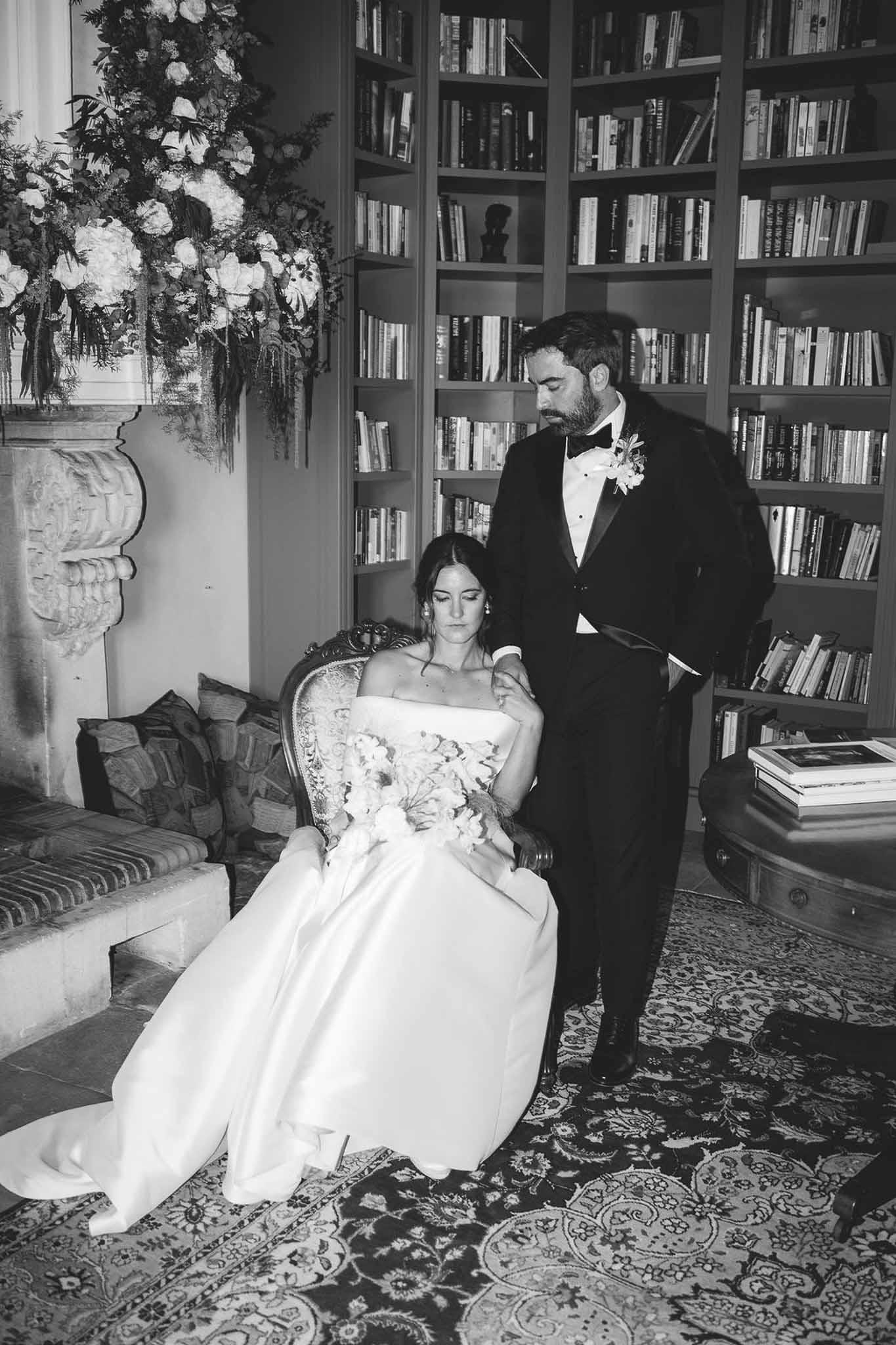 Black and white portrait of bride seated in armchair with groom standing behind in library with bookshelves