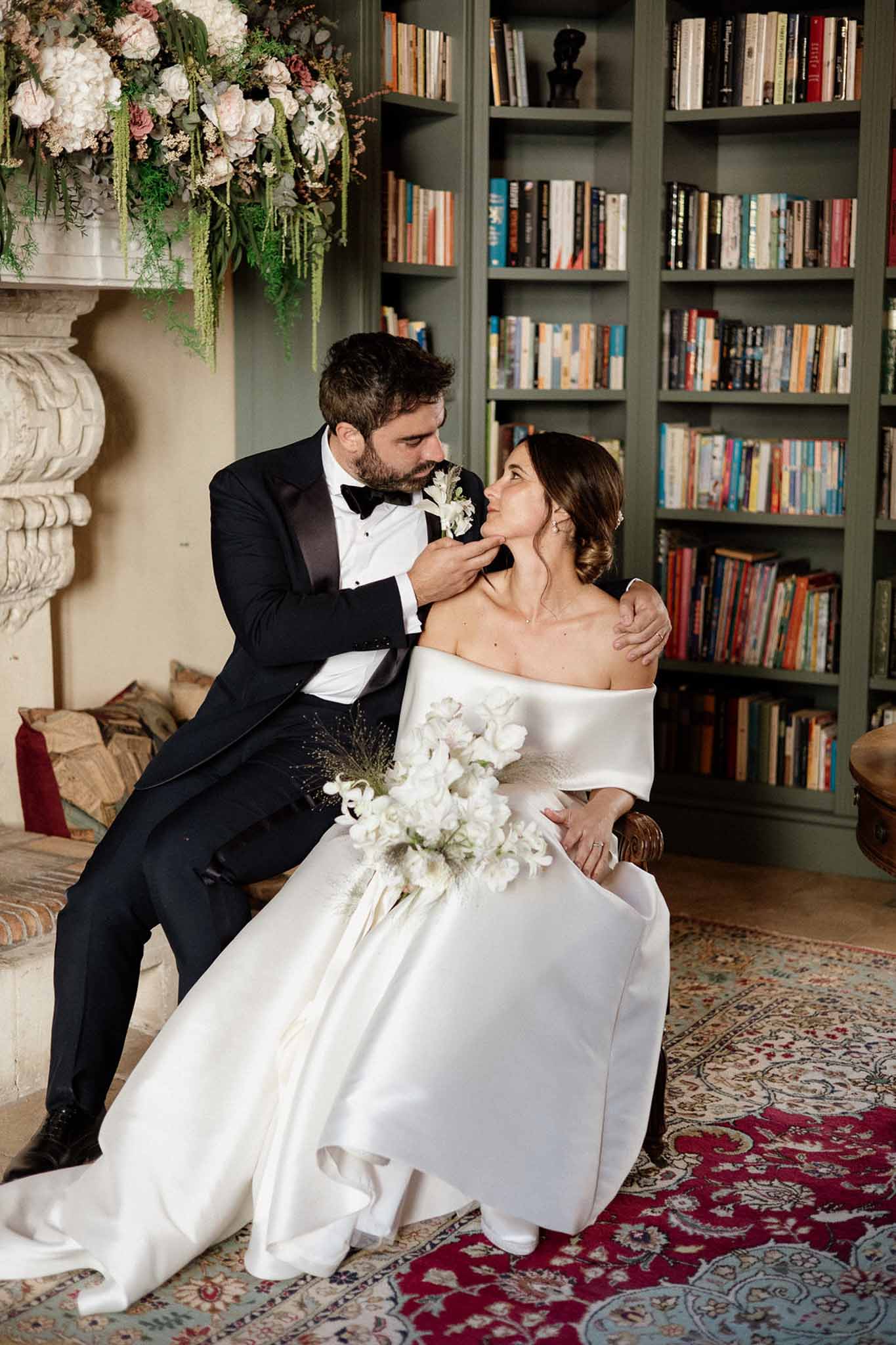 Groom in black tuxedo cupping bride's face while seated by fireplace in library with white floral arrangements