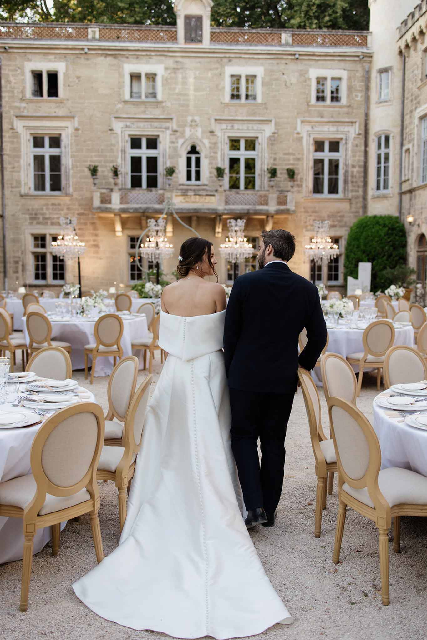 Bride and groom walk through chateau courtyard reception with round tables, chandeliers, and gold chairs
