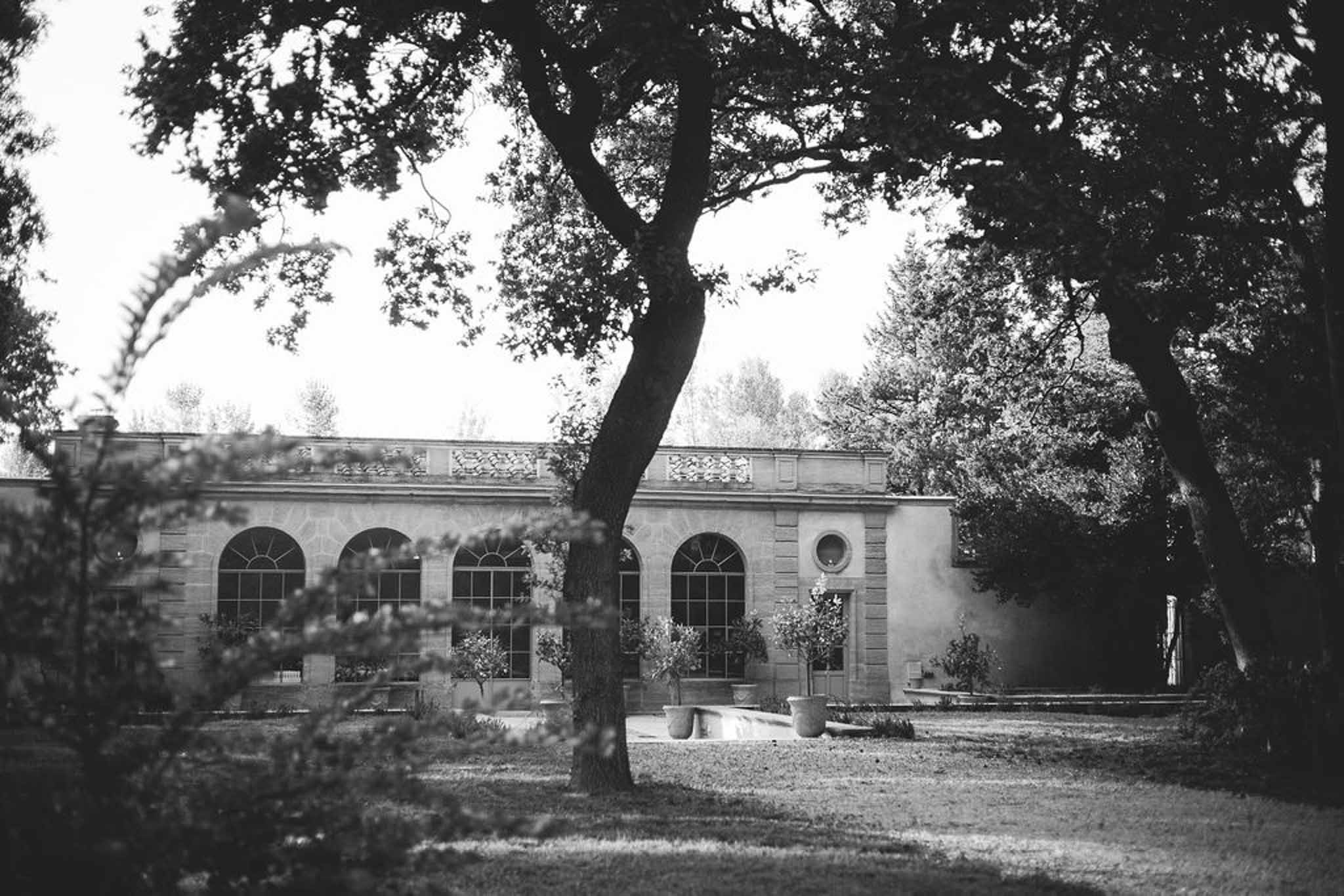 Black and white exterior of arched orangerie with stone friezes and terracotta planters through trees