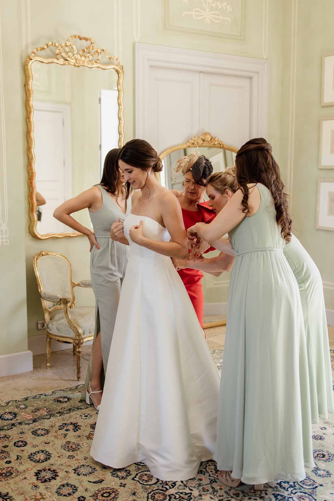 Bride being helped into her strapless gown by bridesmaids in sage green dresses in a chateau bridal suite