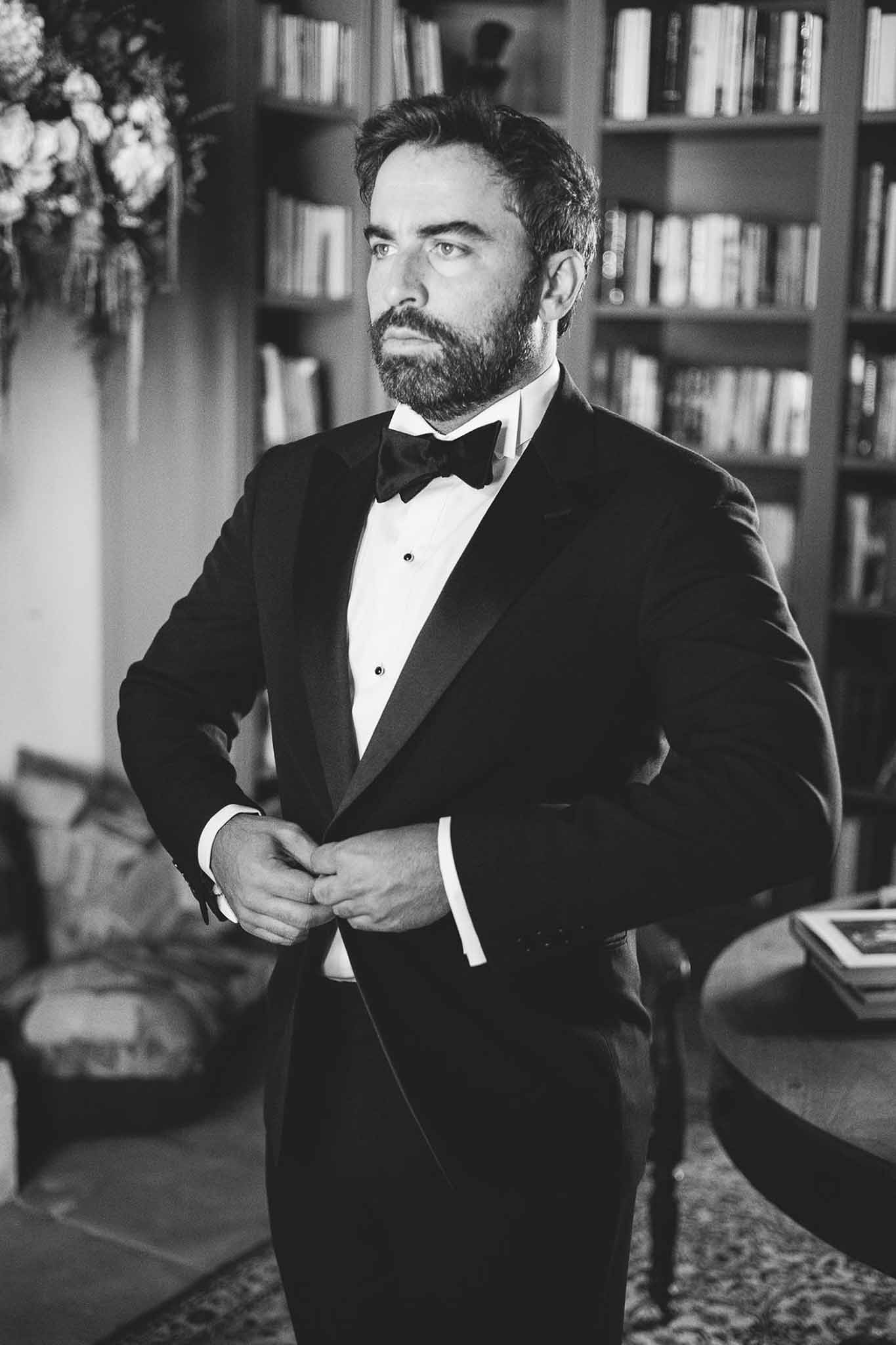 Black-and-white portrait of groom buttoning tuxedo jacket in library with floor-to-ceiling bookshelves